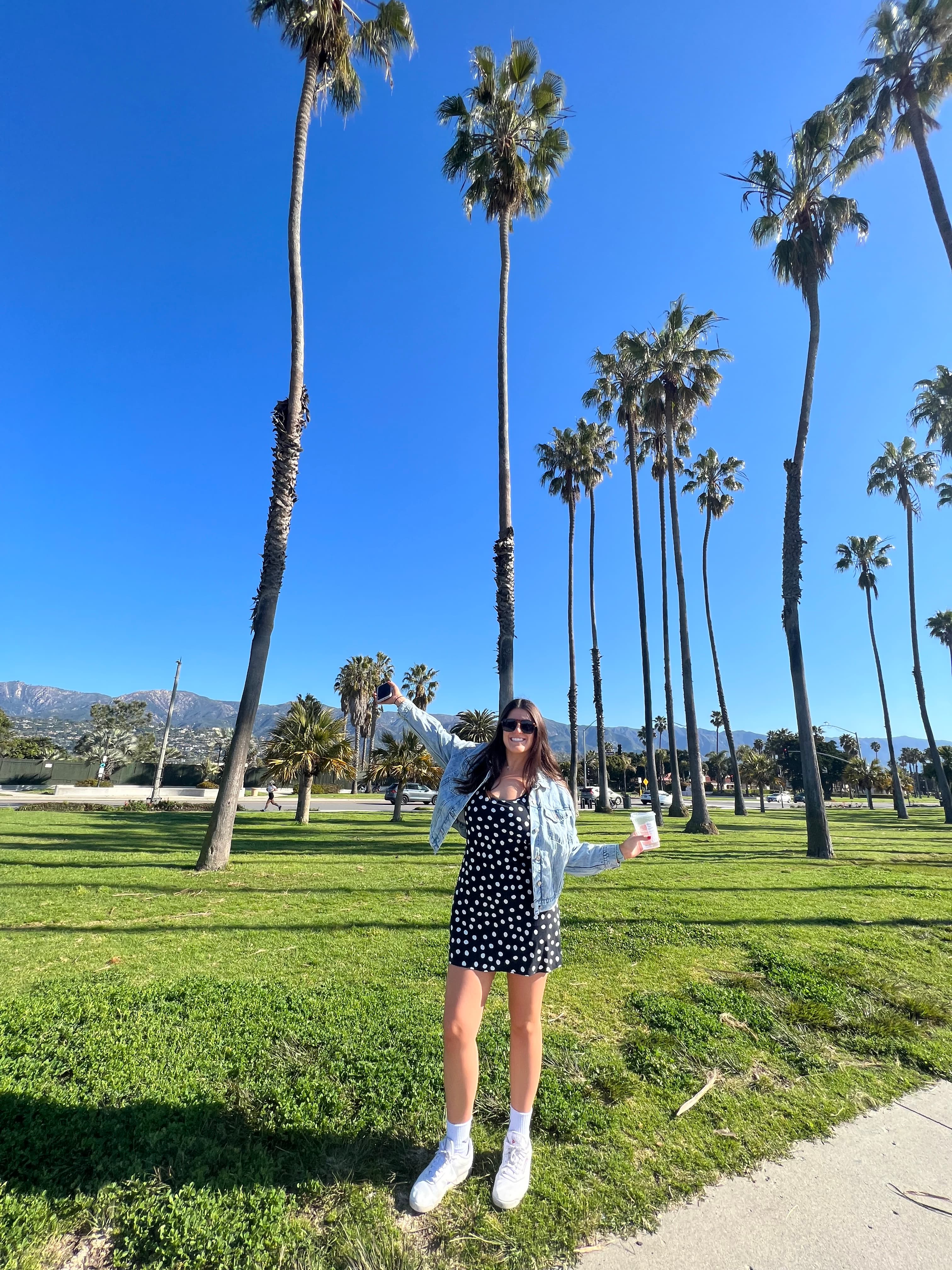 Lauren in a denim jacket posing in a grassy area with several tall palm trees behind her on a sunny day