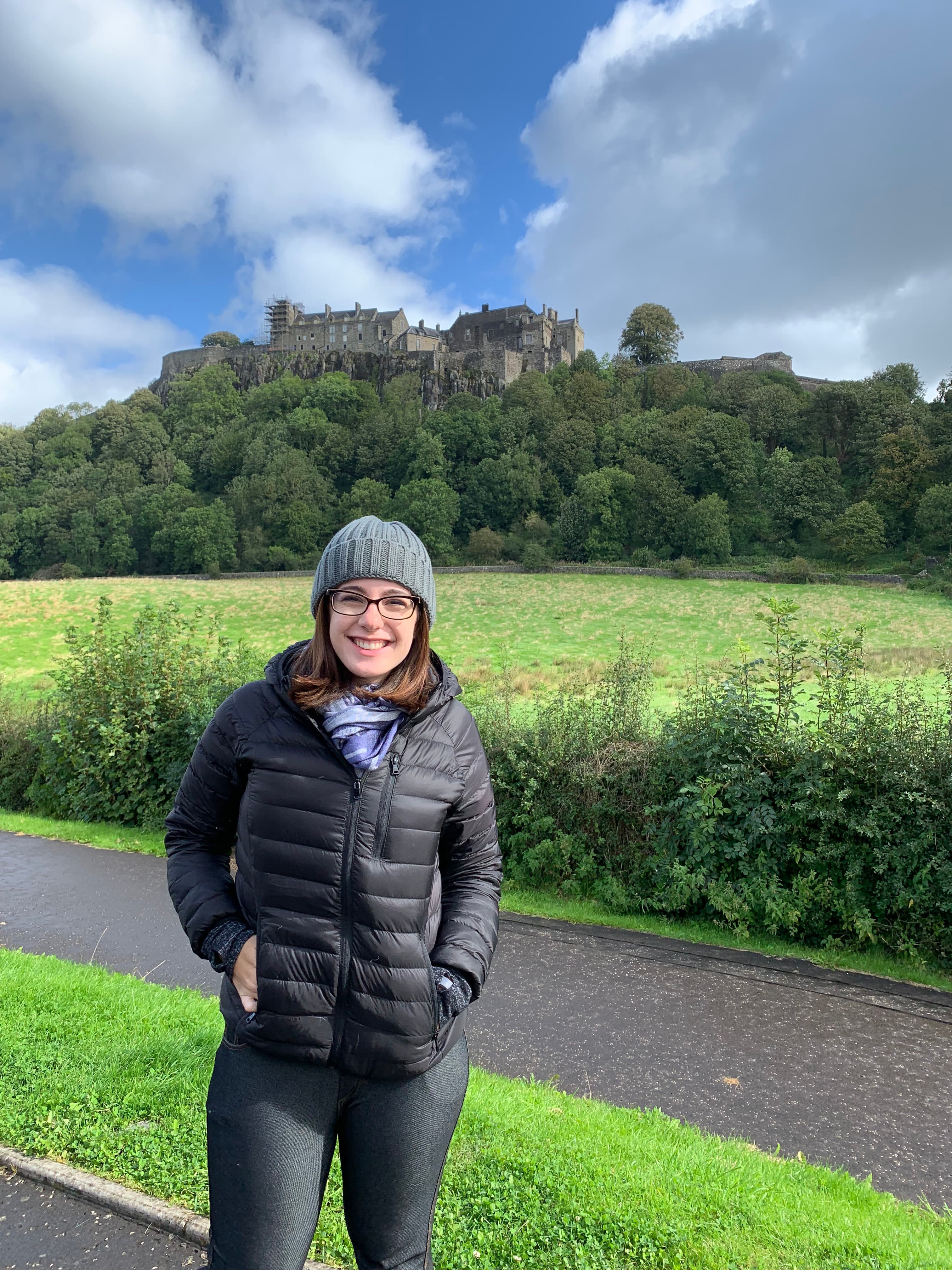 Advisor in a black winter coat and hat posing in front of a wooden area with a castle rising above the trees