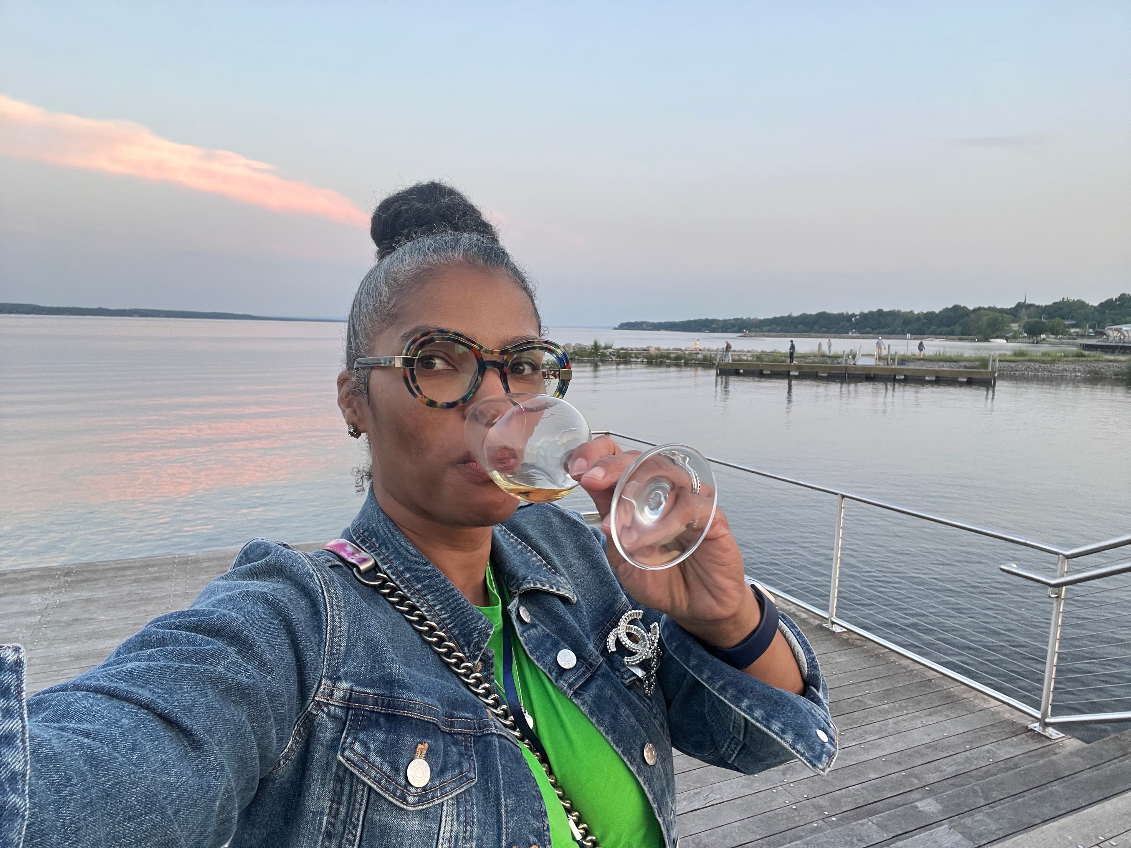 Katrina in a denim jacket sipping from a glass of wine while taking a selfie overlooking the sea