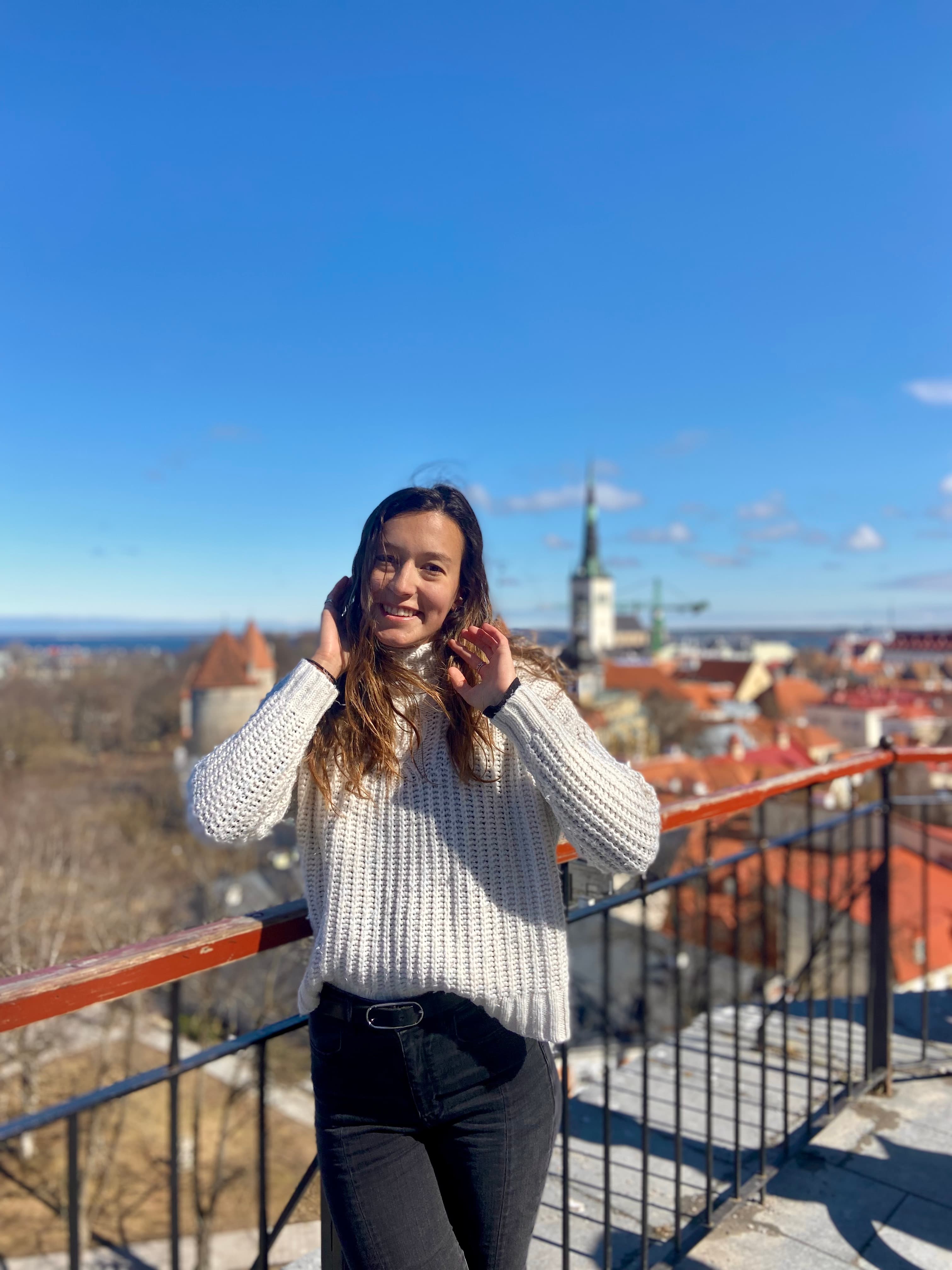 Advisor in a grey sweater posing on a rooftop overlooking orange tiled buildings and the sea visible on the horizon