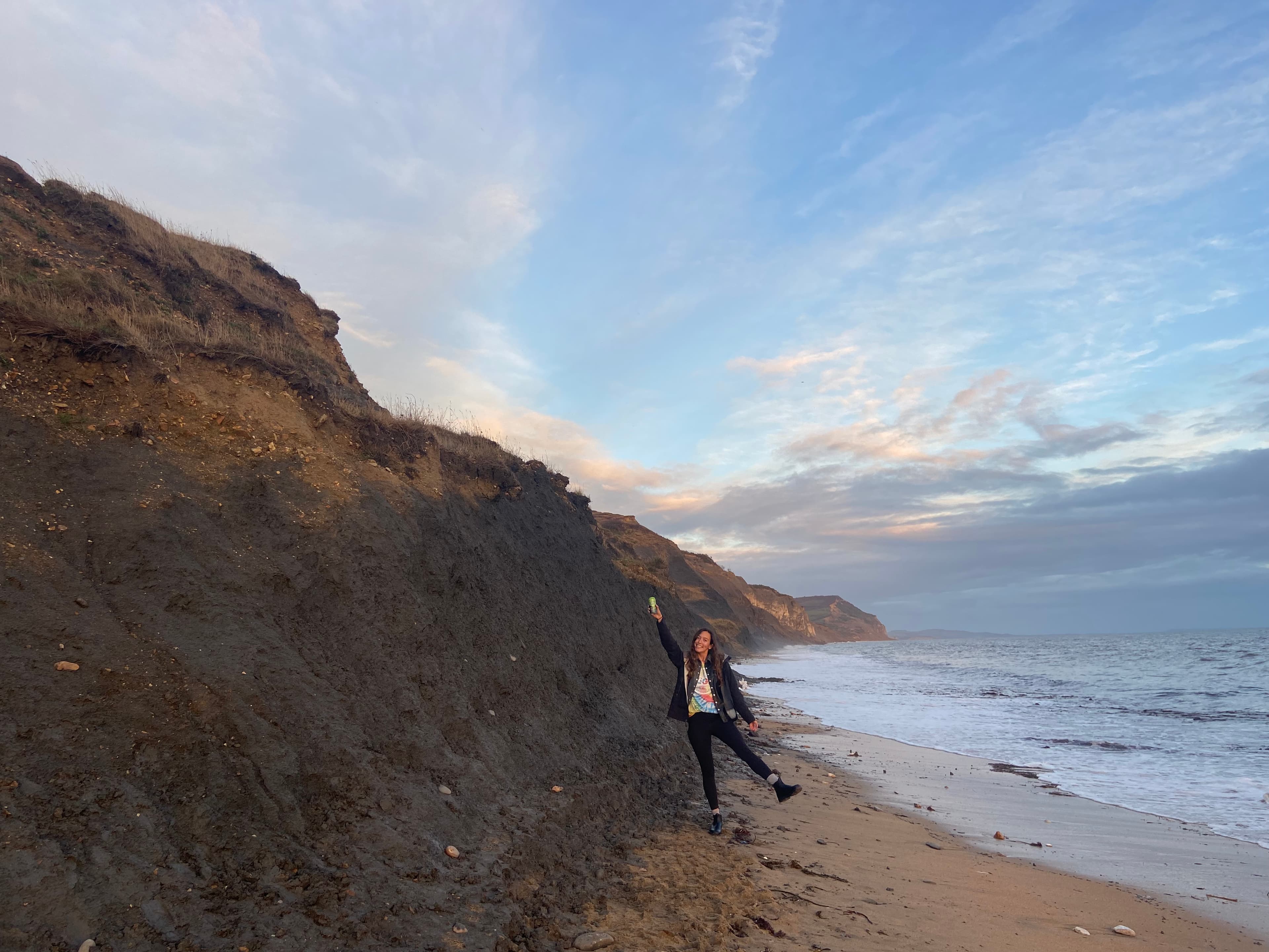 Advisor standing on one foot with her arm raised on a beach with rocky cliffs