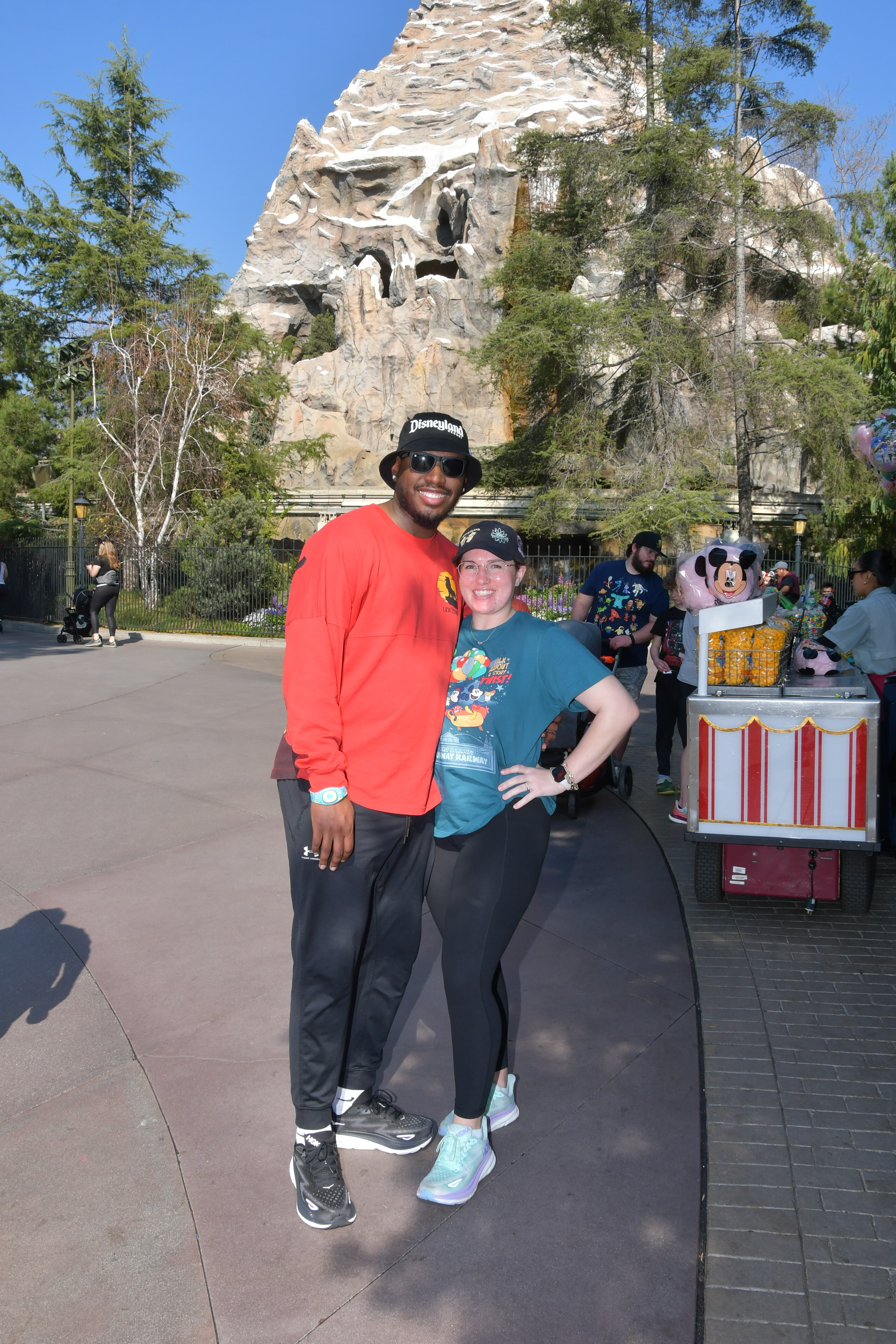 Staci and a man posing side by side outdoors with trees and a food vendor behind them