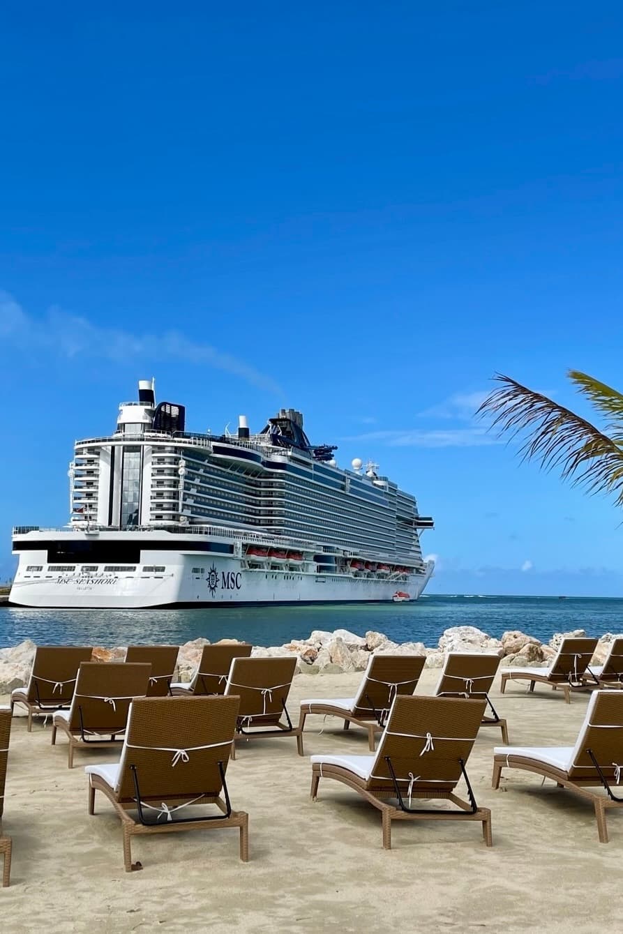 A view of a cruise ship docked at a port with beach loungers on a sunny day. 