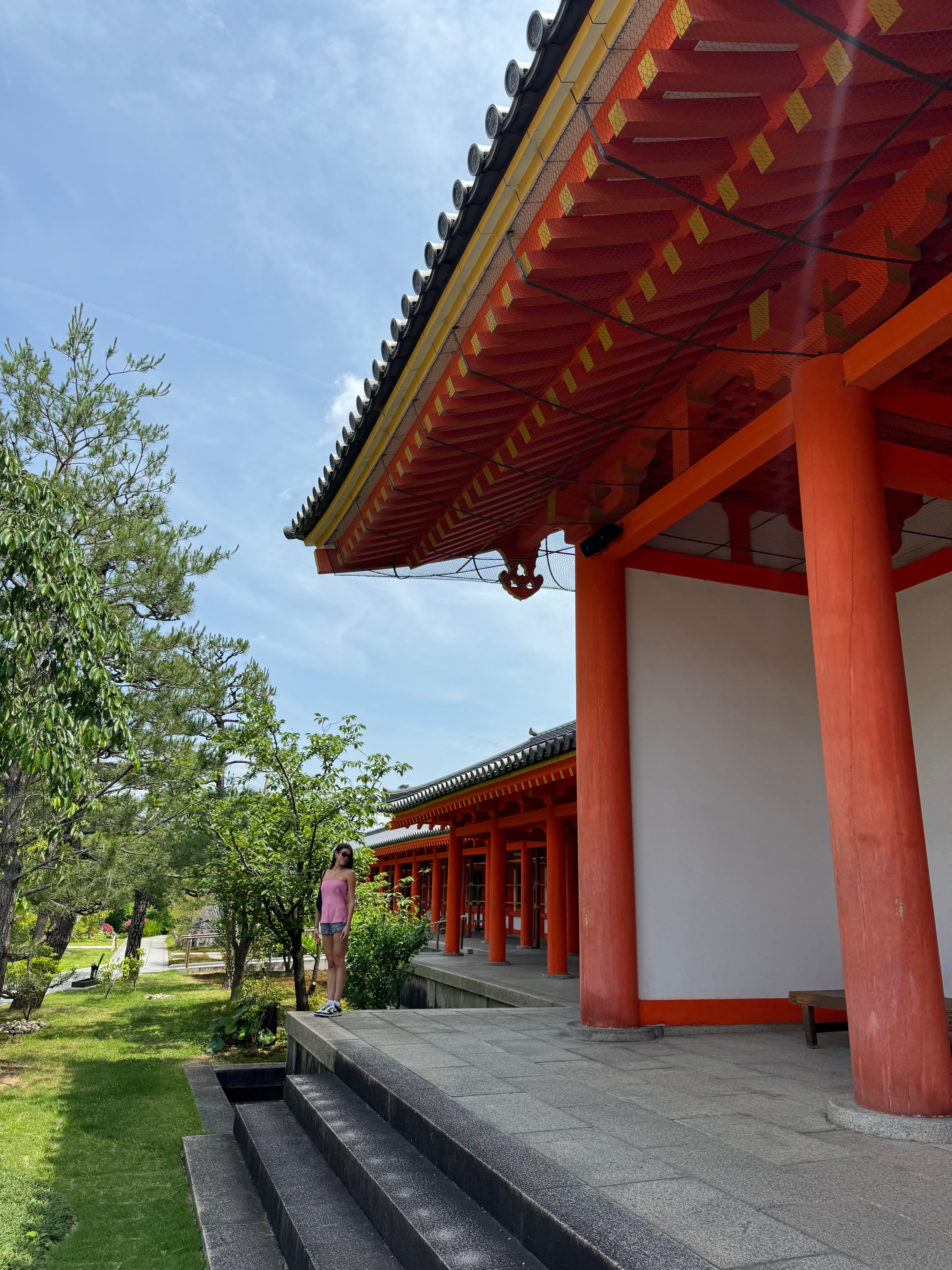 View of the side of a red Japanese temple and nearby garden area on a sunny day