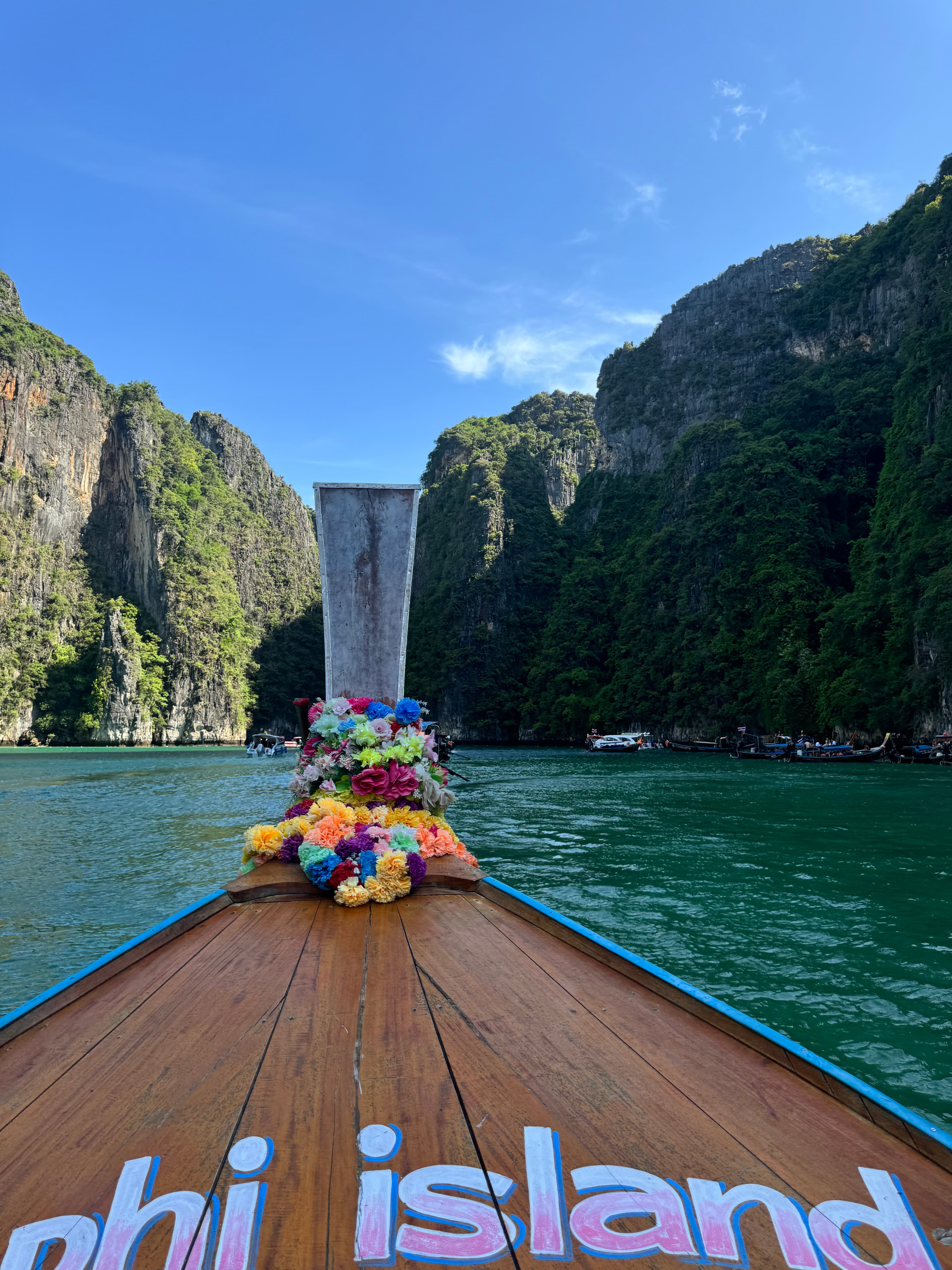 View of the front of a boat reading “PHI ISLAND” with flowers at the front sailing by stunning green cliffs