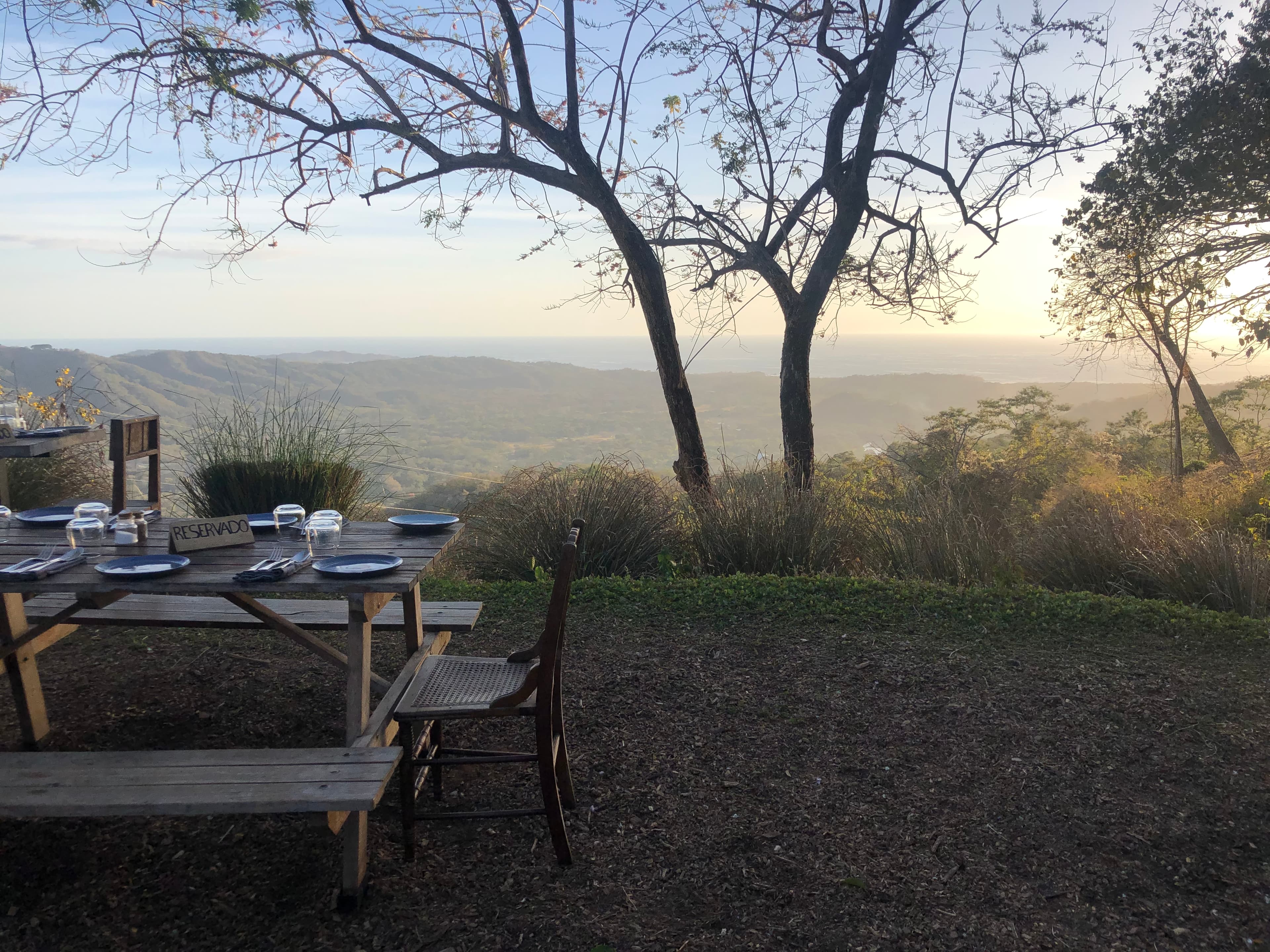 Beautiful view of a wooden picnic table set with plates overlooking a valley at sunset