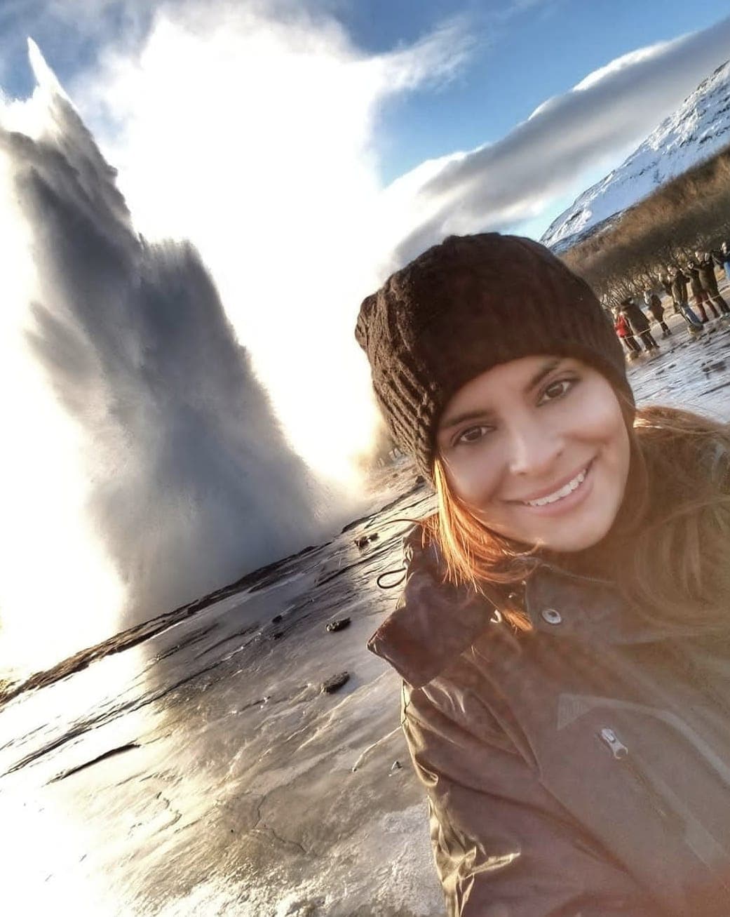 Melissa in a black beanie taking a selfie in front of an erupting geyser on a sunny day