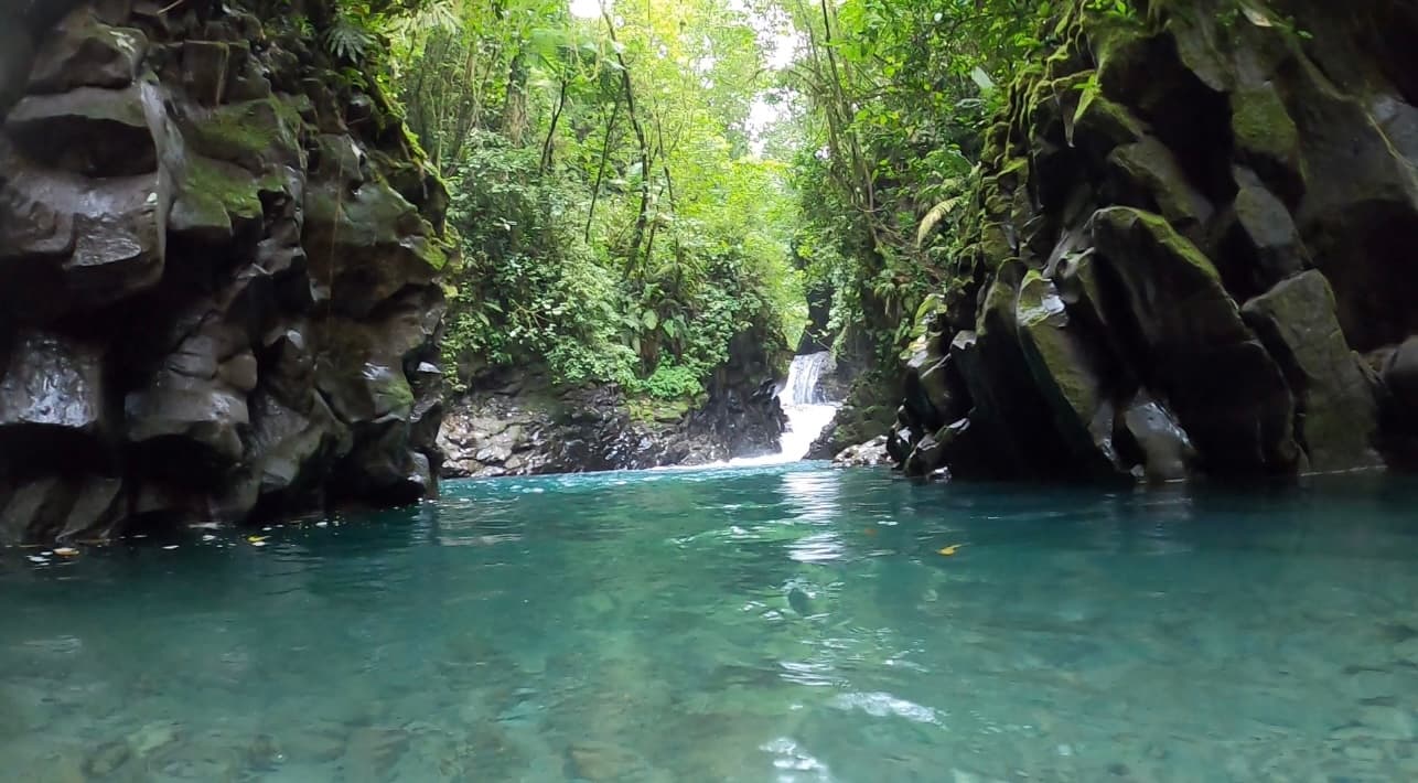 An image of a body of water between two rock formations leading out to a lush forest area and waterfall.