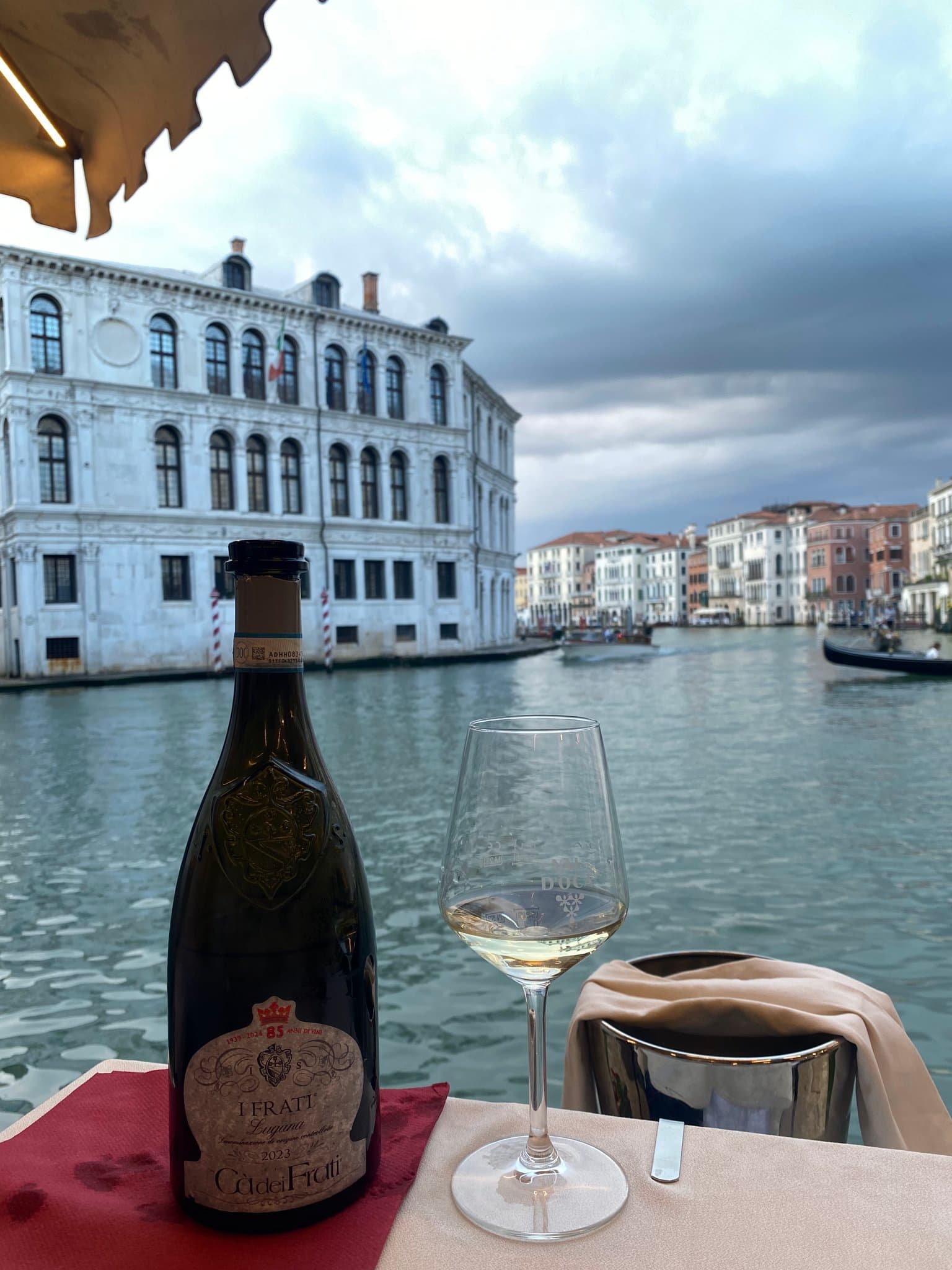A bottle of wine and glass at the edge of a table overlooking the water's edge on a clear day.