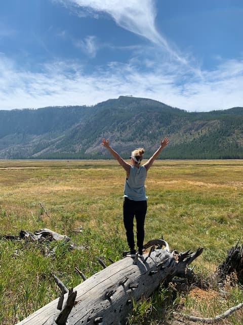 Advisor standing on a fallen tree trunk with arms raised in the middle of a vast field, mountains visible in the distance