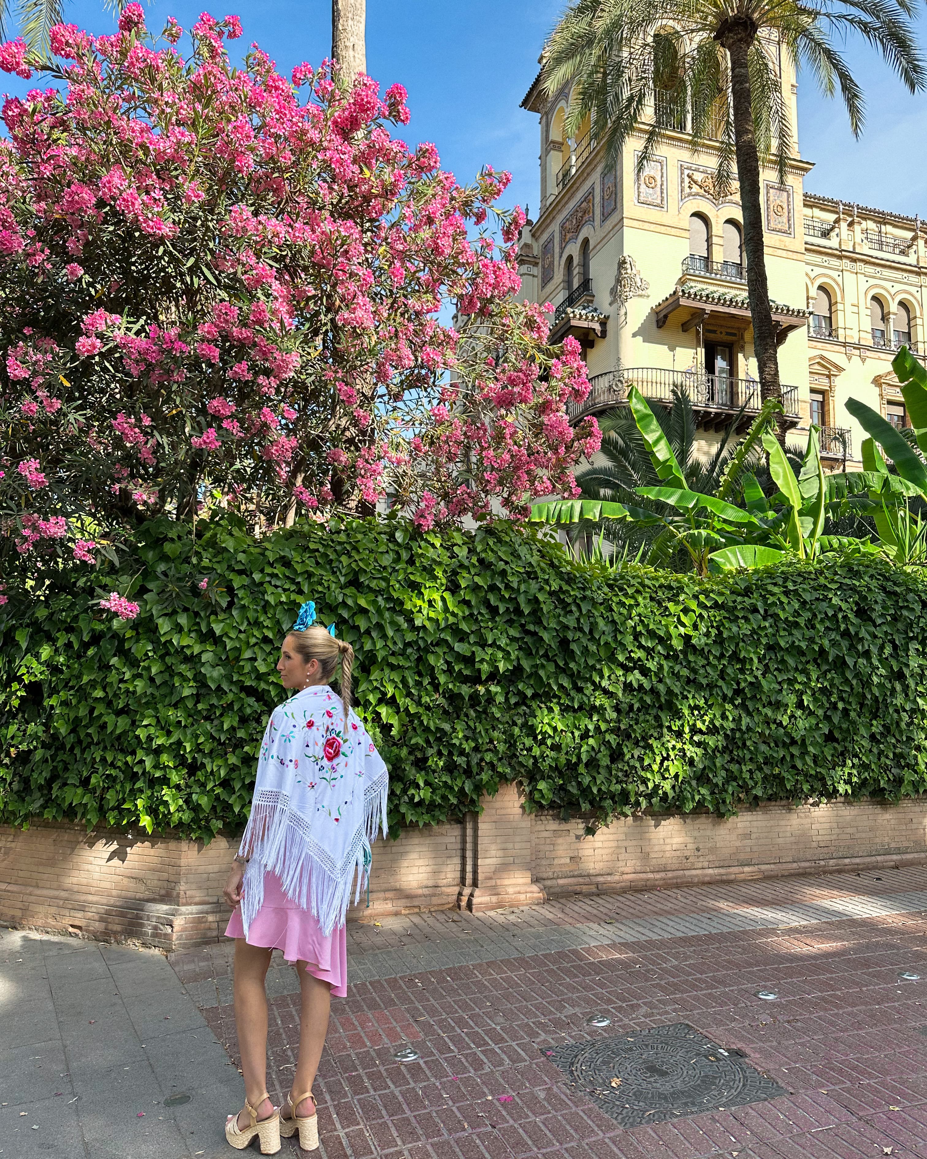 A woman dressed in a pink and white outfit and looking out to a bush and pink tree. There is a palm tree and beautiful building in the upper right hand corner.