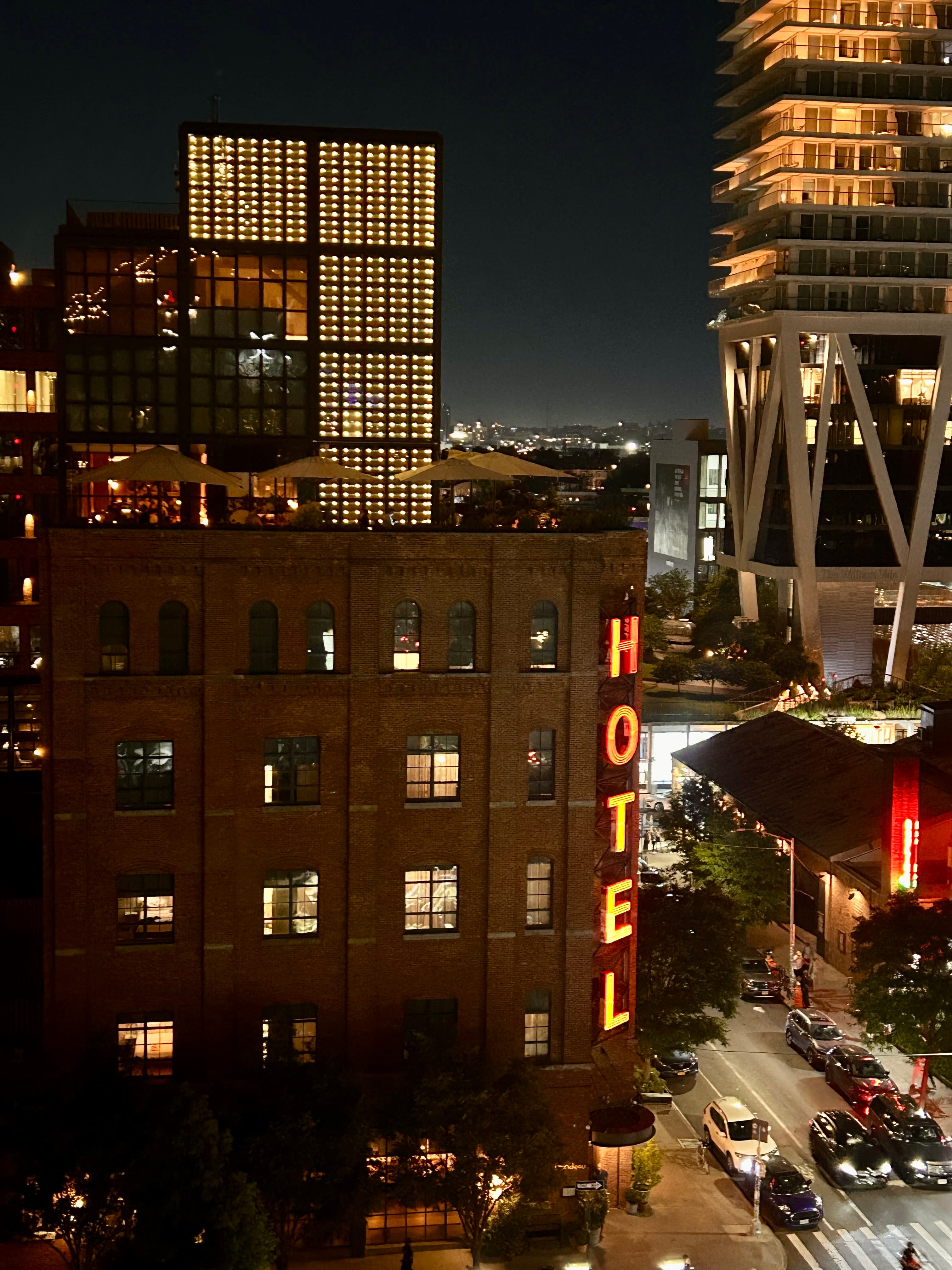 View of a square hotel building on a city corner at night, cars on the street visible below
