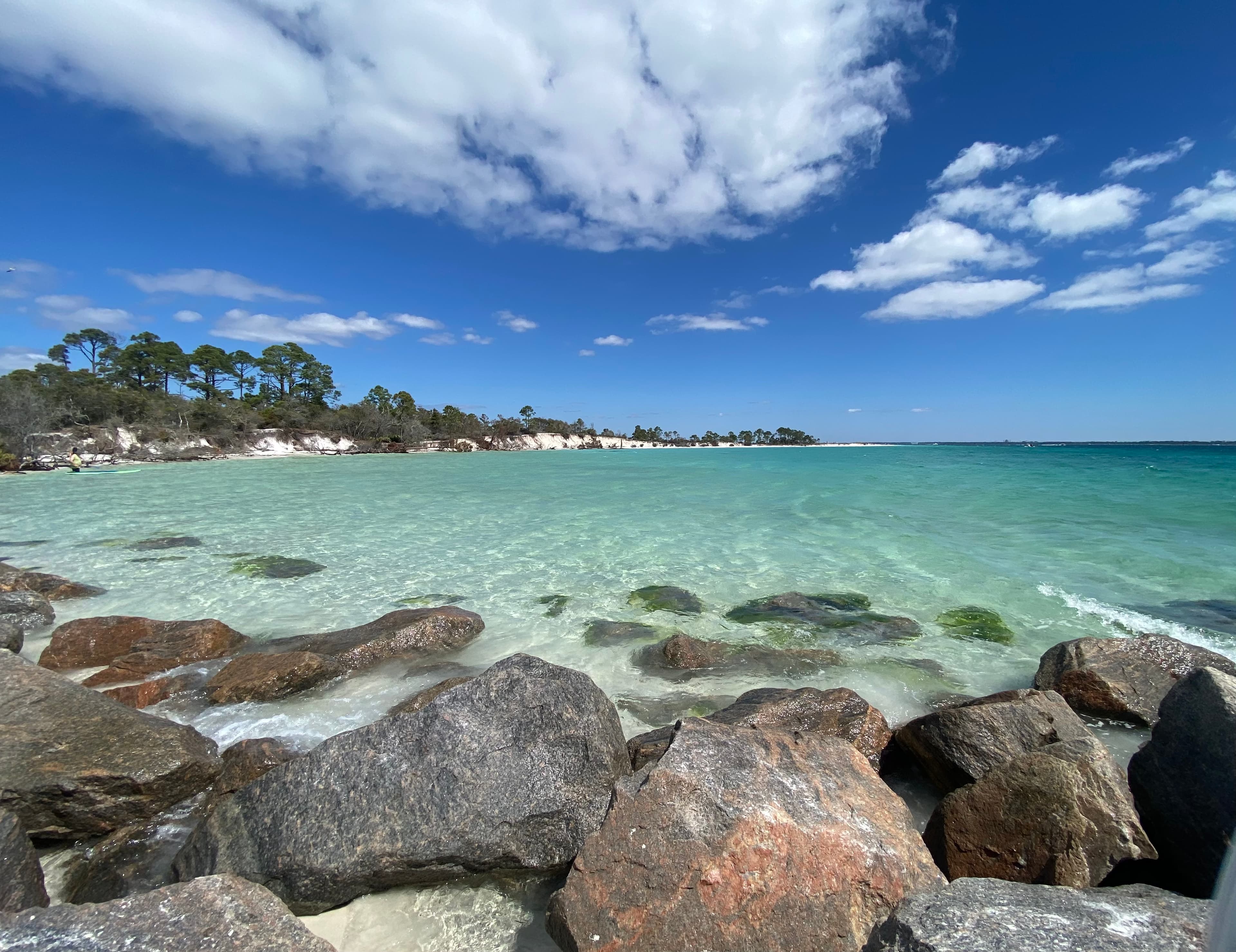 A rocky shoreline with clear blue water and clouds overhead.