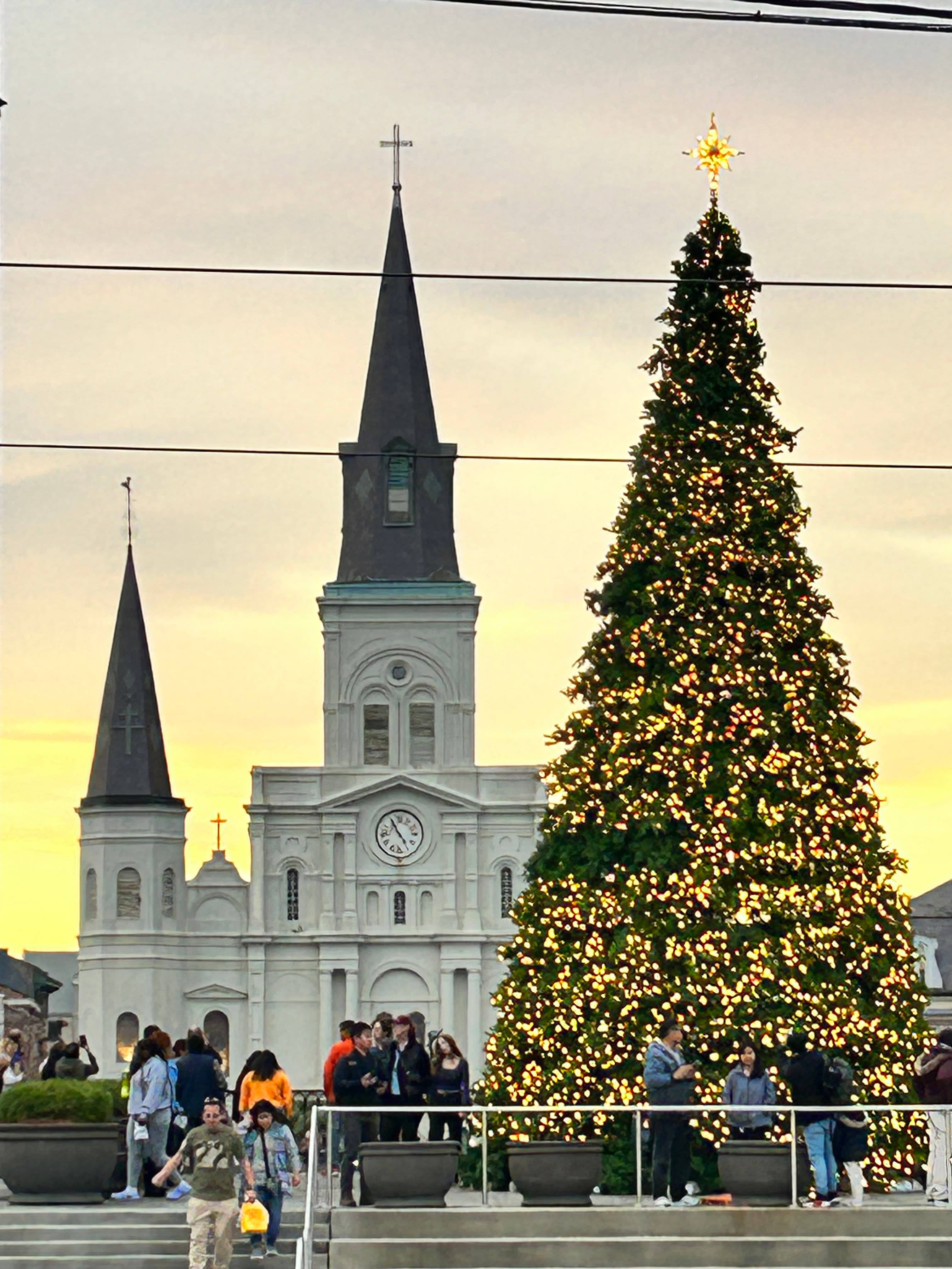 A view of a tall, decorated Christmas tree with a church and sunset in the background. 