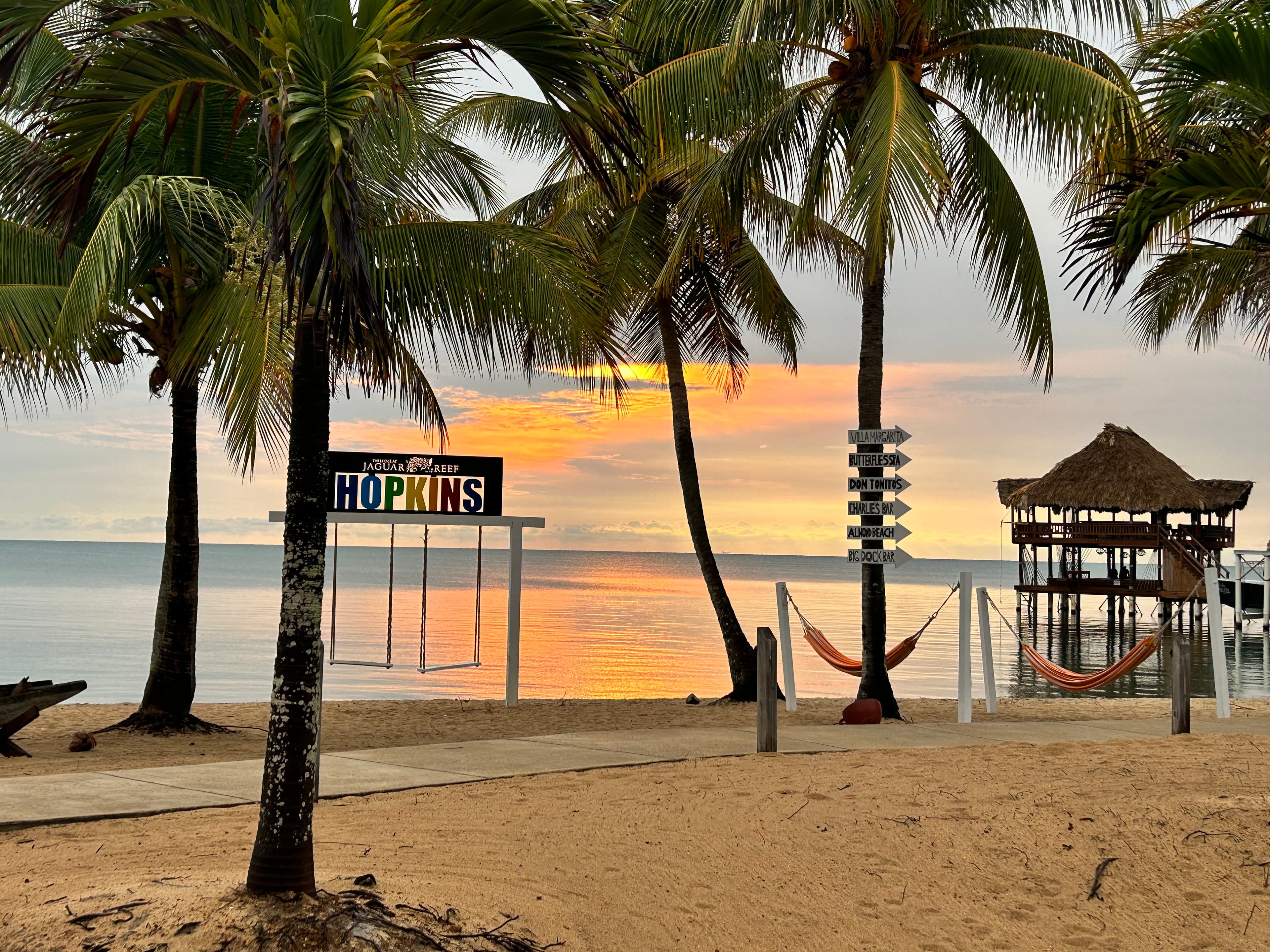 A view of the beach with palm trees, hammocks, cabanas, and a golden, orange sunset shining over the water. 