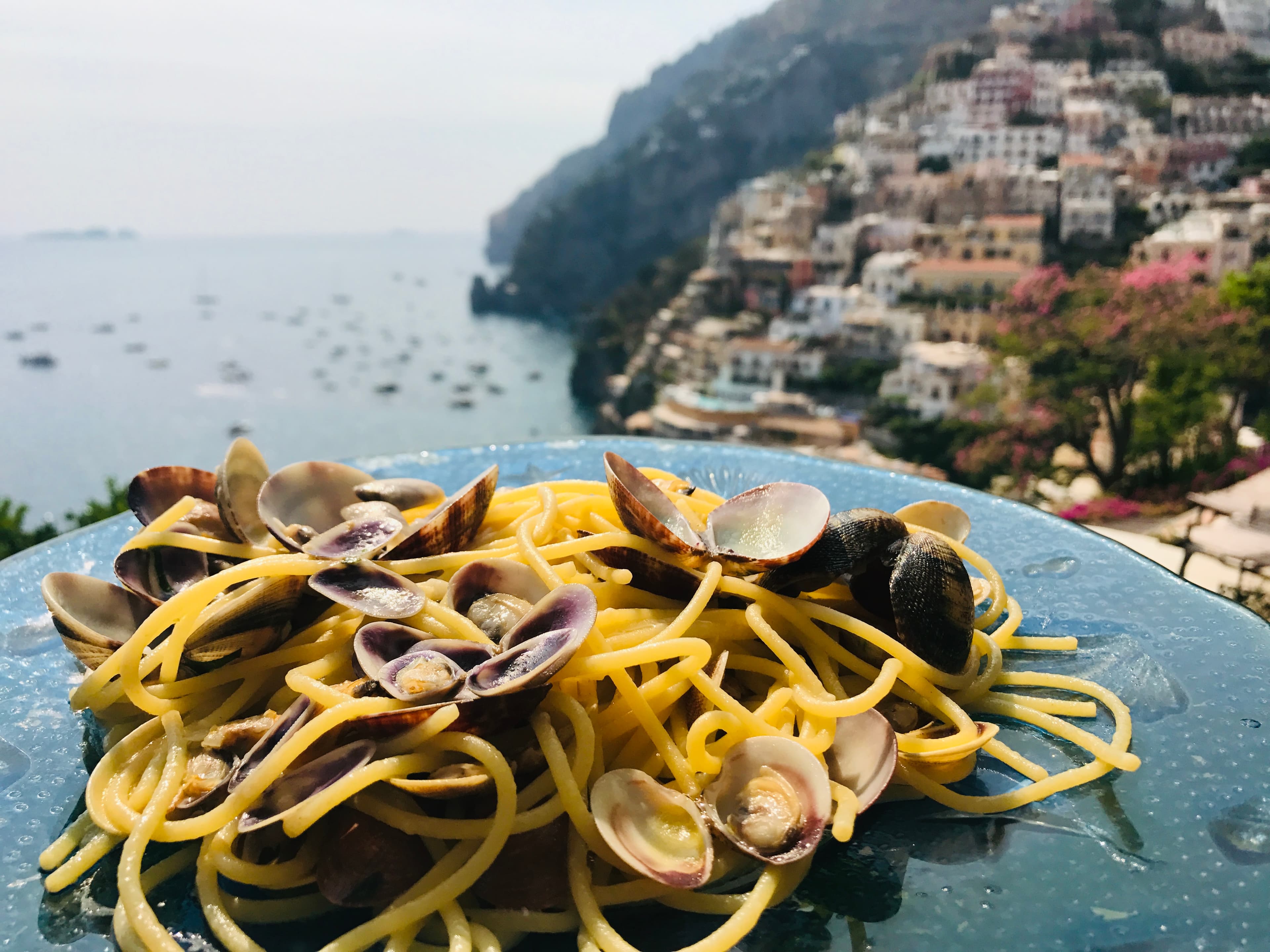 View of a plate of spaghetti with clams overlooking a small Italian coastal city with boats floating offshore