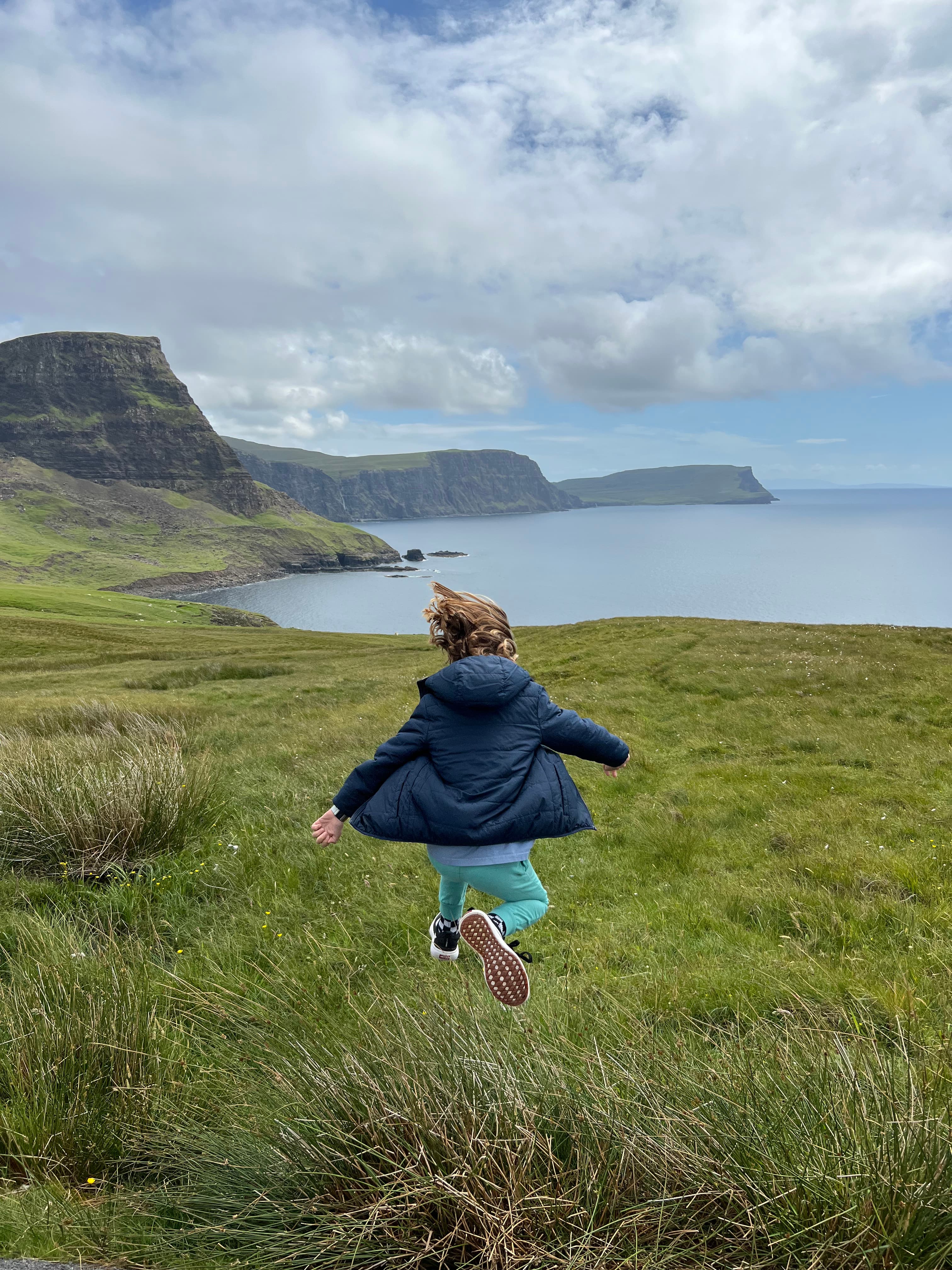 A child runs through a grassy field with sea cliffs in the distance.
