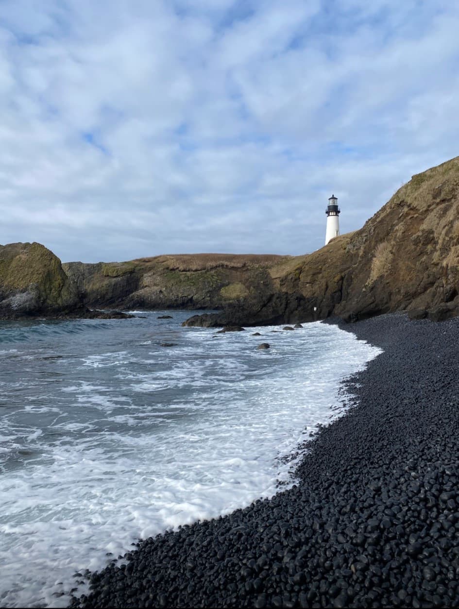 A lighthouse stands tall against a cloudy sky as waves crash against the black sand beach below. 