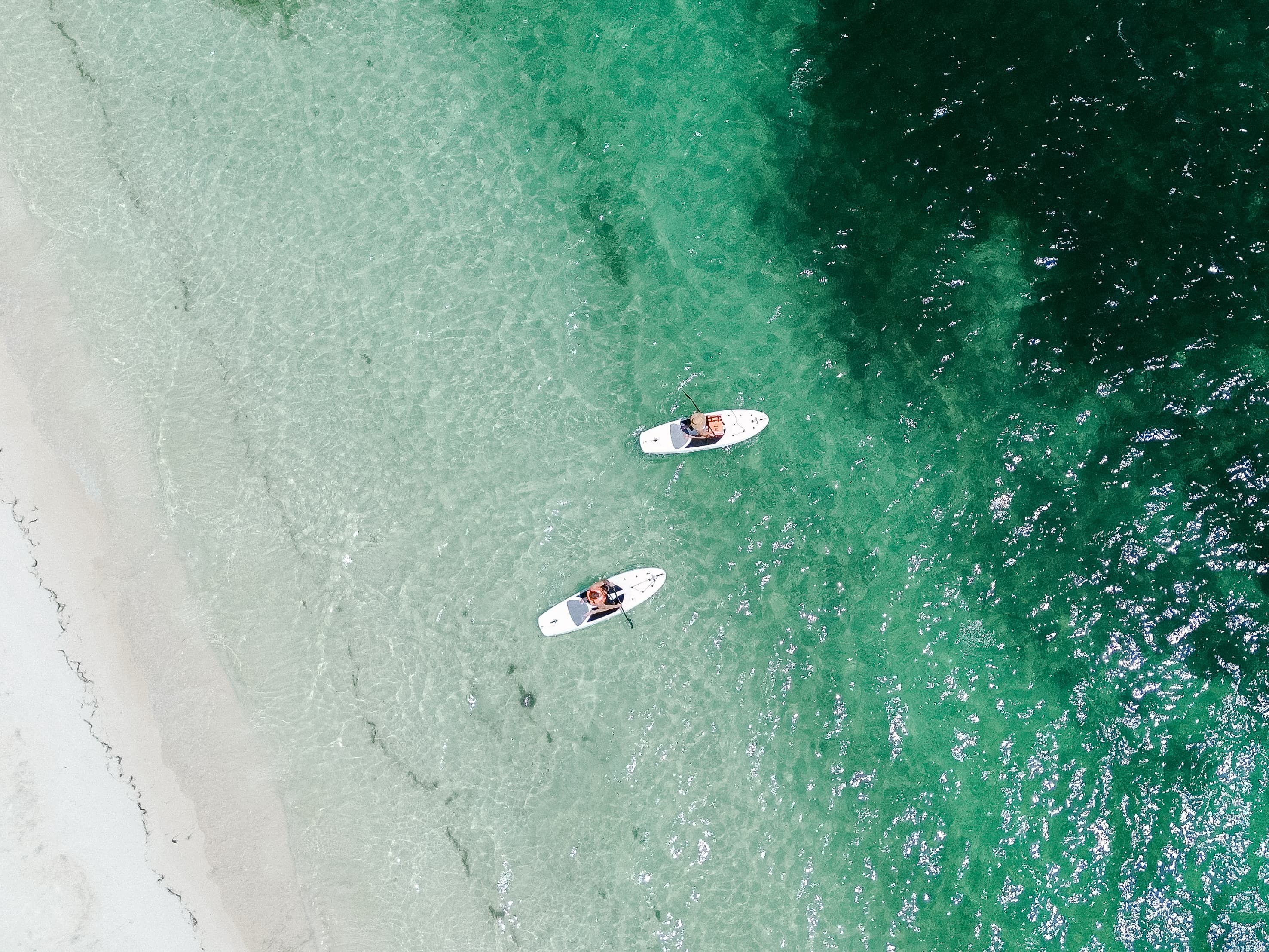 An aerial view of a couple sitting on top of white paddle boards floating over crystal-clear ocean waters. 