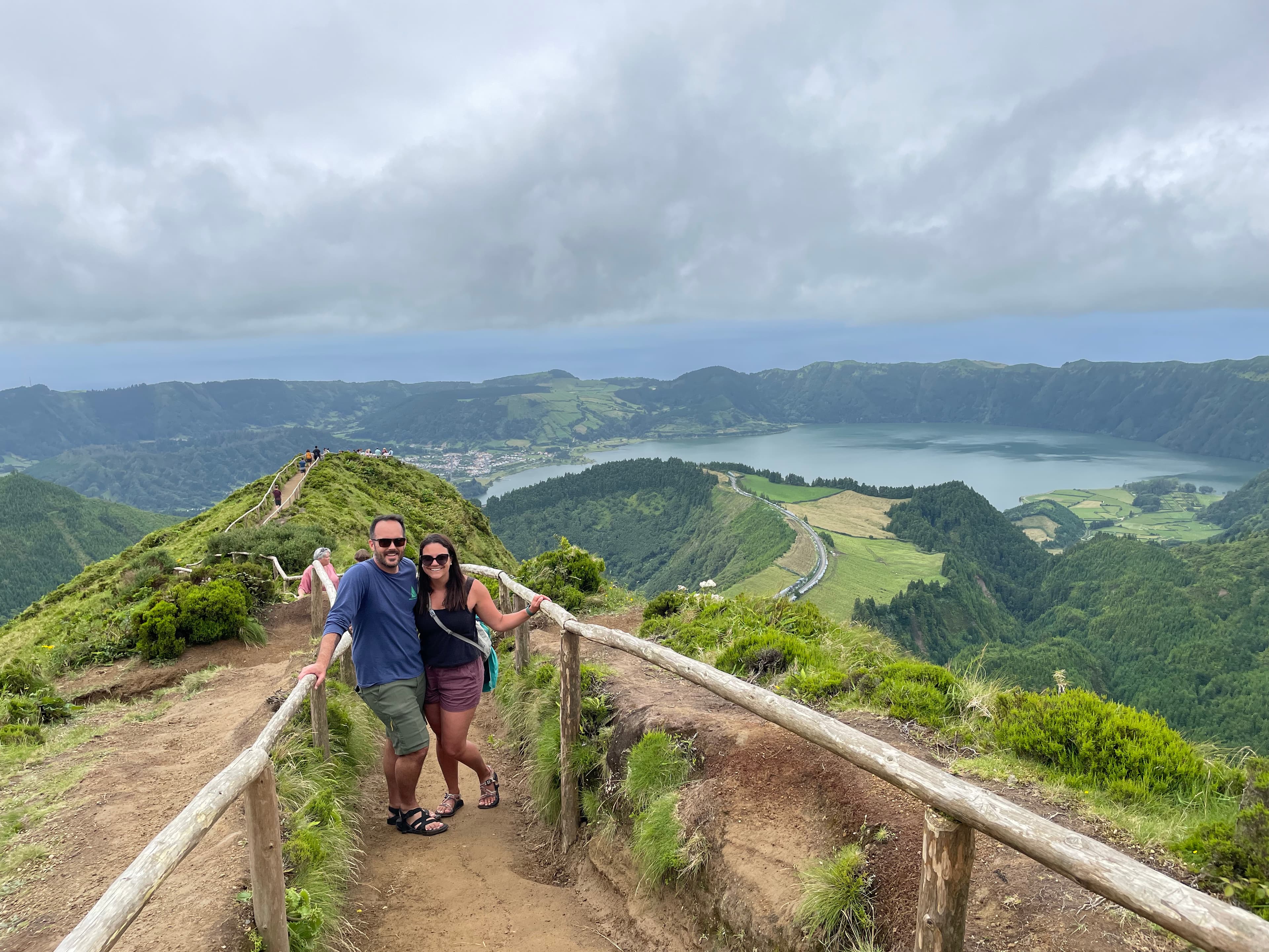 Two people enjoy the scenic view from a wooden walkway atop a mountain, surrounded by vast landscapes.