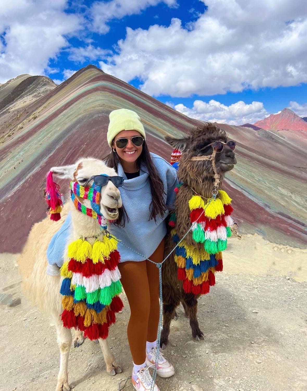 Caity poses with a llama on each side of her on a mountain, surrounded by stunning natural scenery and clear blue skies.