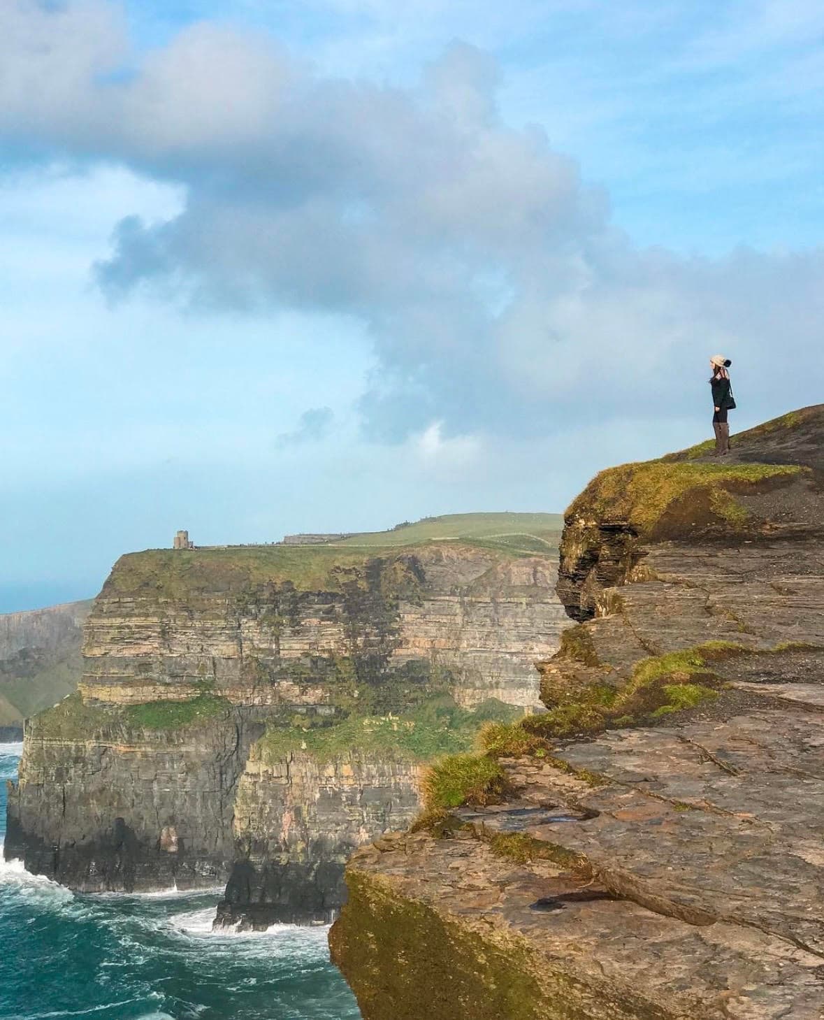 A person stands at the cliff's edge, gazing out over the vast, serene ocean below, with waves crashing against the rocks.
