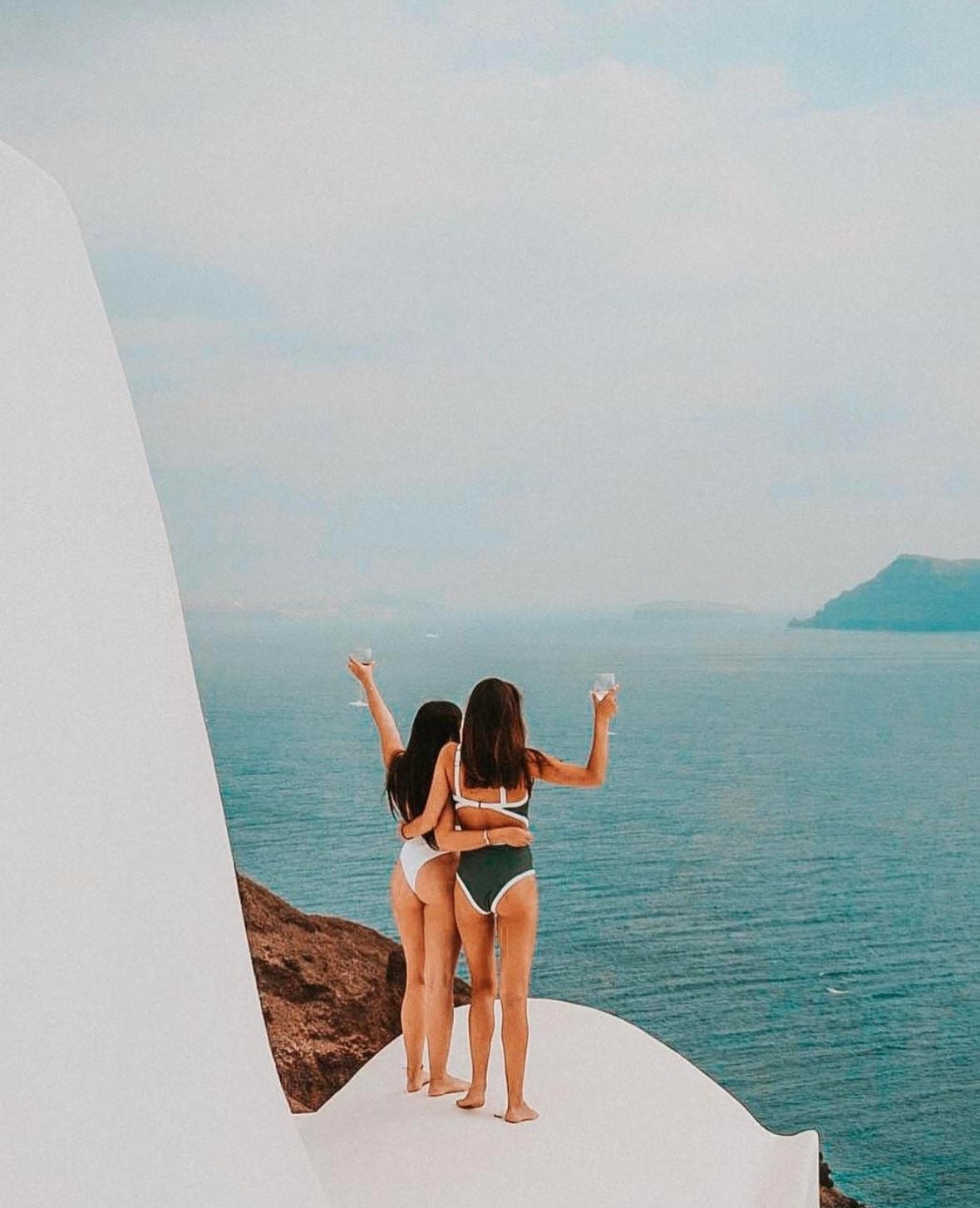 Two women in bikinis stand atop a cliff, admiring the ocean view holding drinks in the air.
