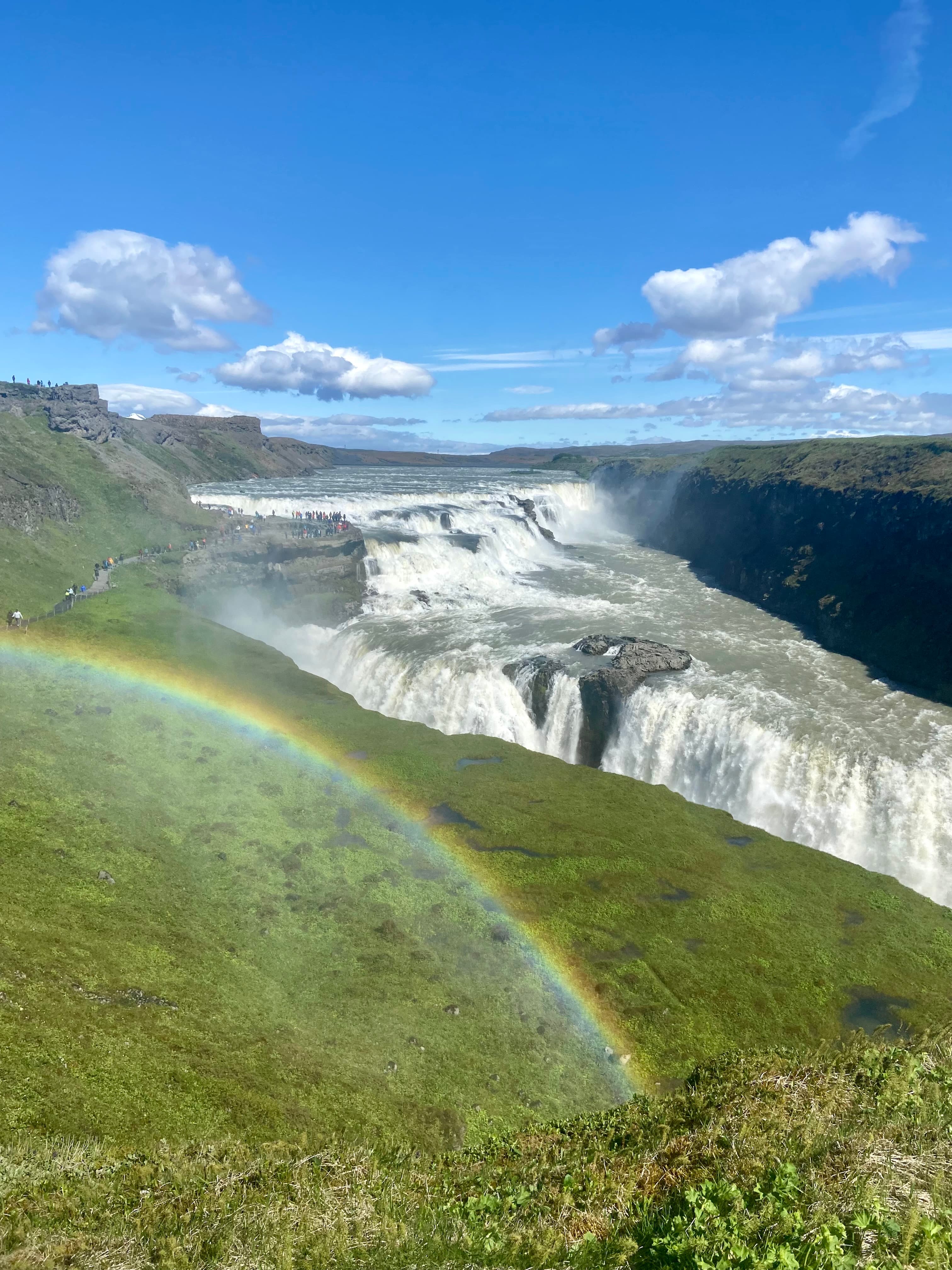 A view of waterfalls and a rainbow shining above them, while surrounded by grassy rolling hills. 