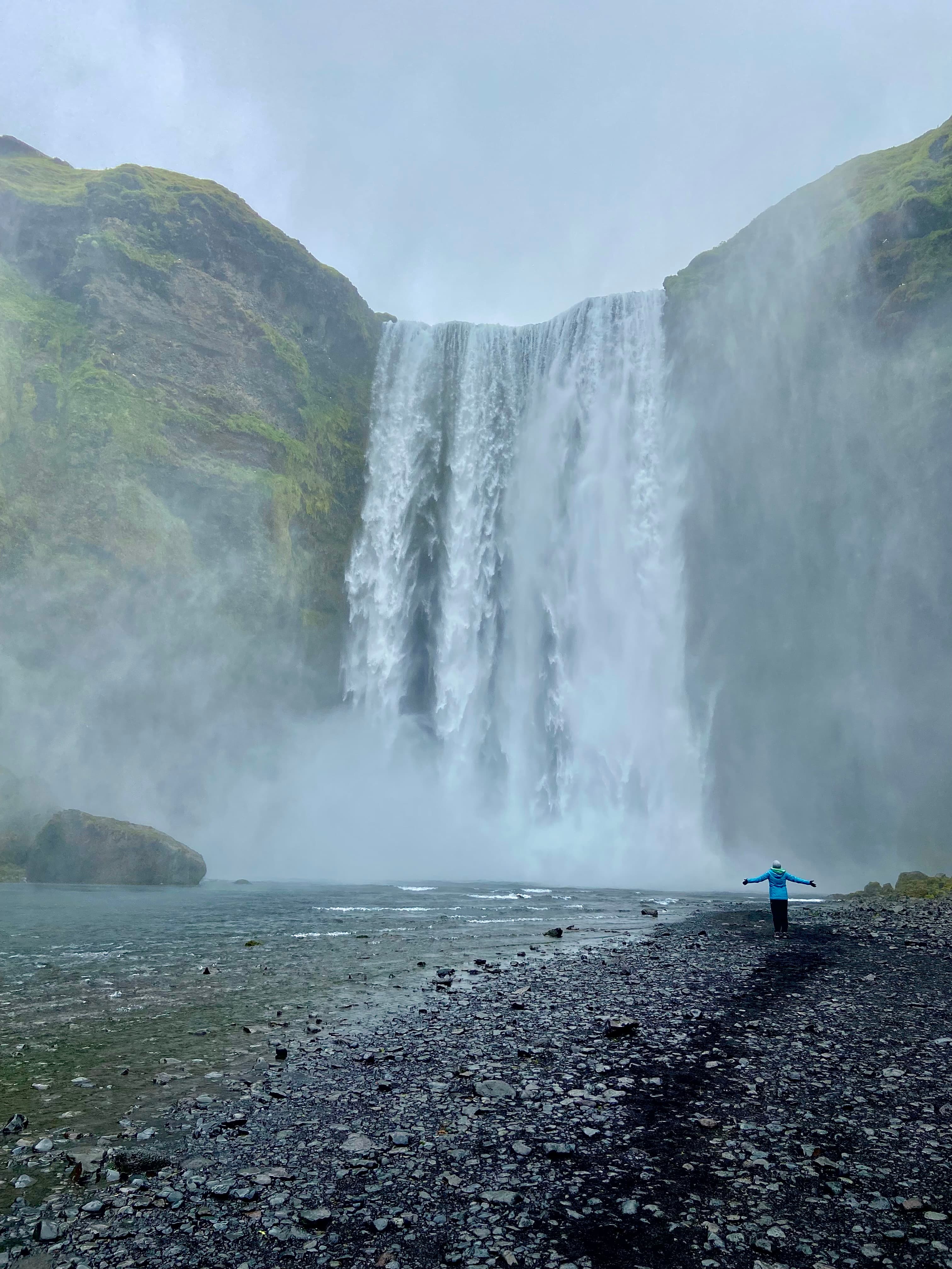 A large waterfall rapidly bellowing downward while a rocky beach and person wearing a blue jacket with their arms outstretched wide awaits at the bottom. 