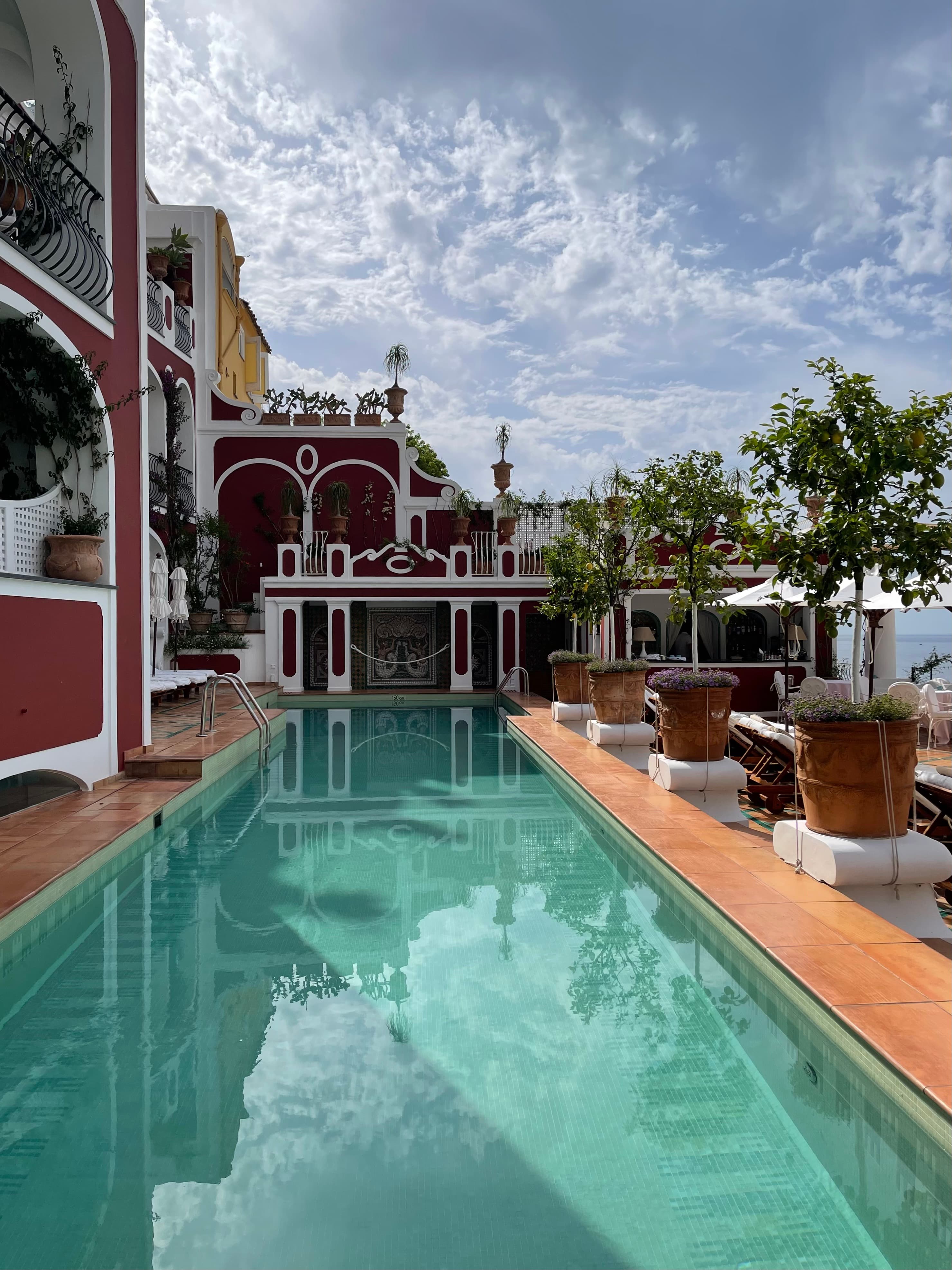 View of a long empty hotel swimming pool next to a red building with white trim on a sunny day