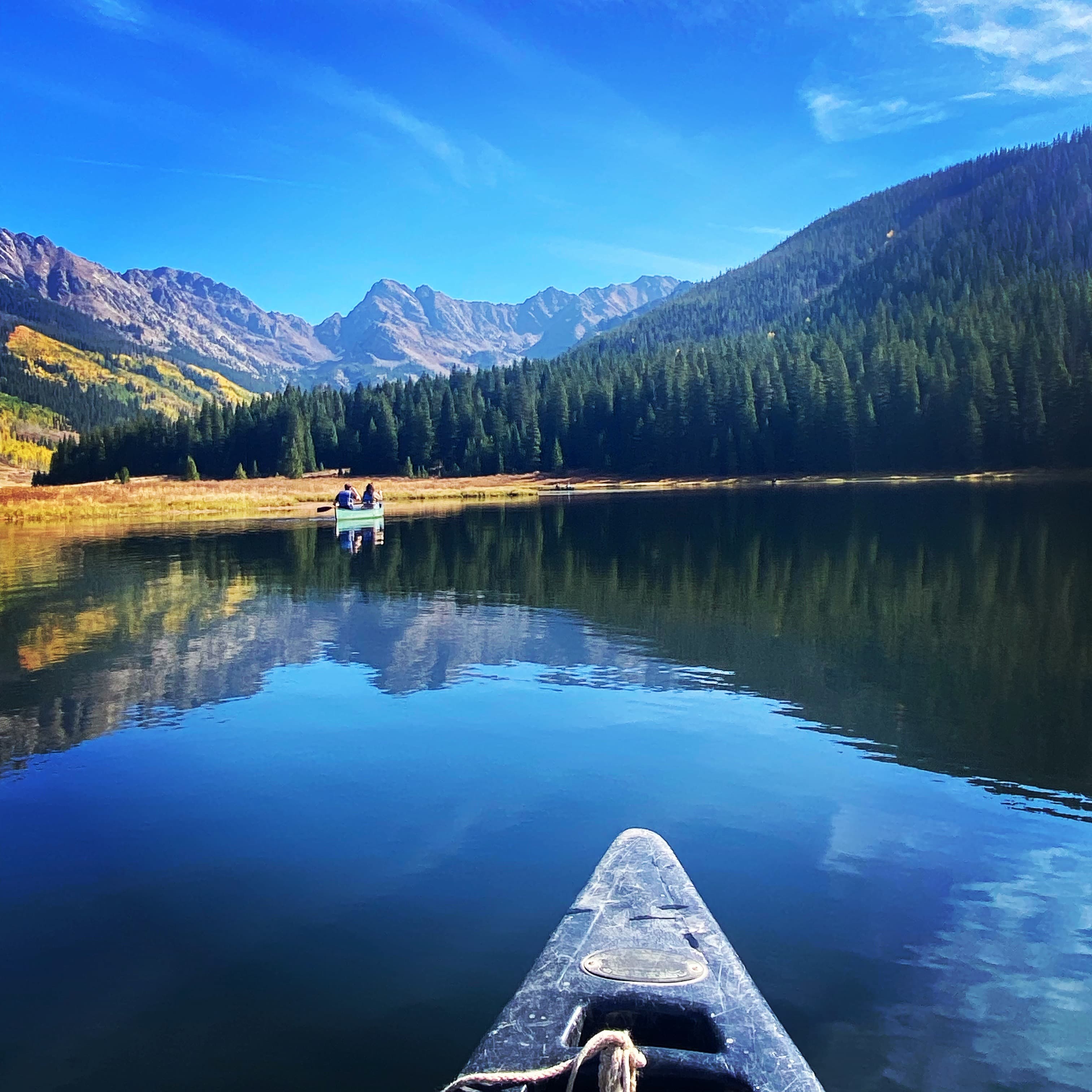View of the front of a kayak on a beautiful glassy lake surrounded by pine trees and stunning mountains