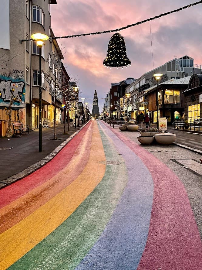 A vibrant rainbow-colored street leads to a distant church, lined with shops and festive decorations, including a large hanging bell under a pink and purple sunset sky.
