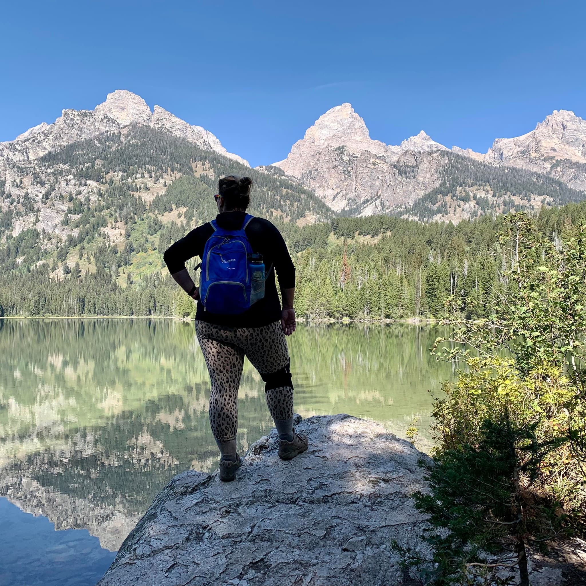 Kate wearing a backpack stands on a rock, gazing at a serene lake surrounded by lush greenery.