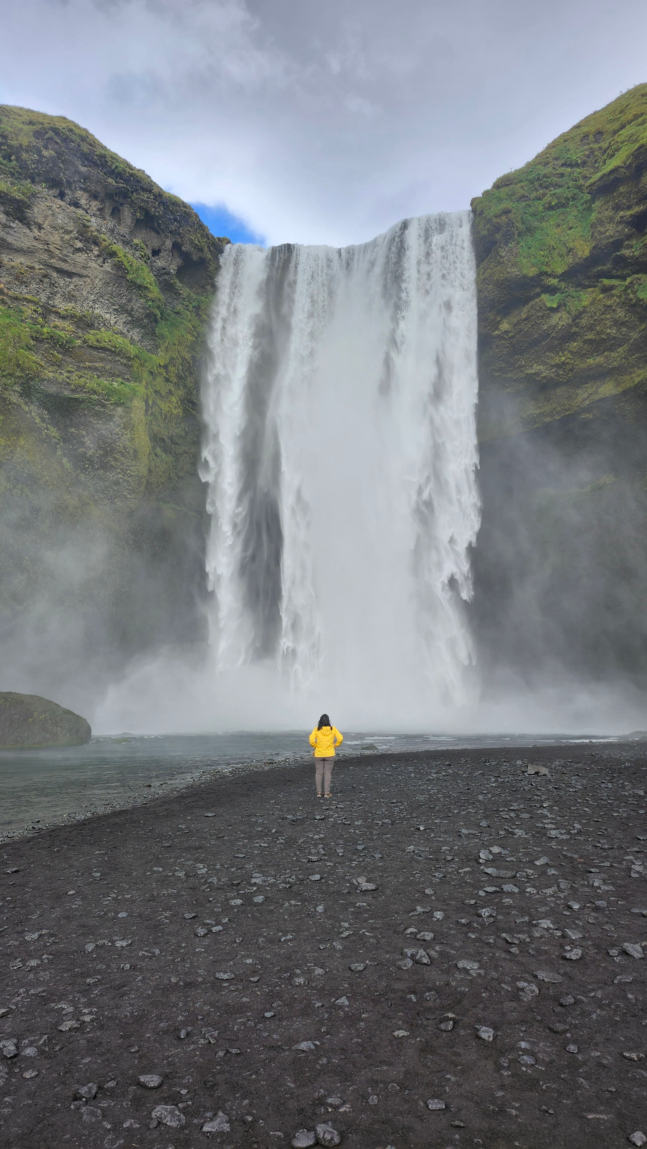 Advisor standing on black rocks in front of a large waterfall on a cloudy day