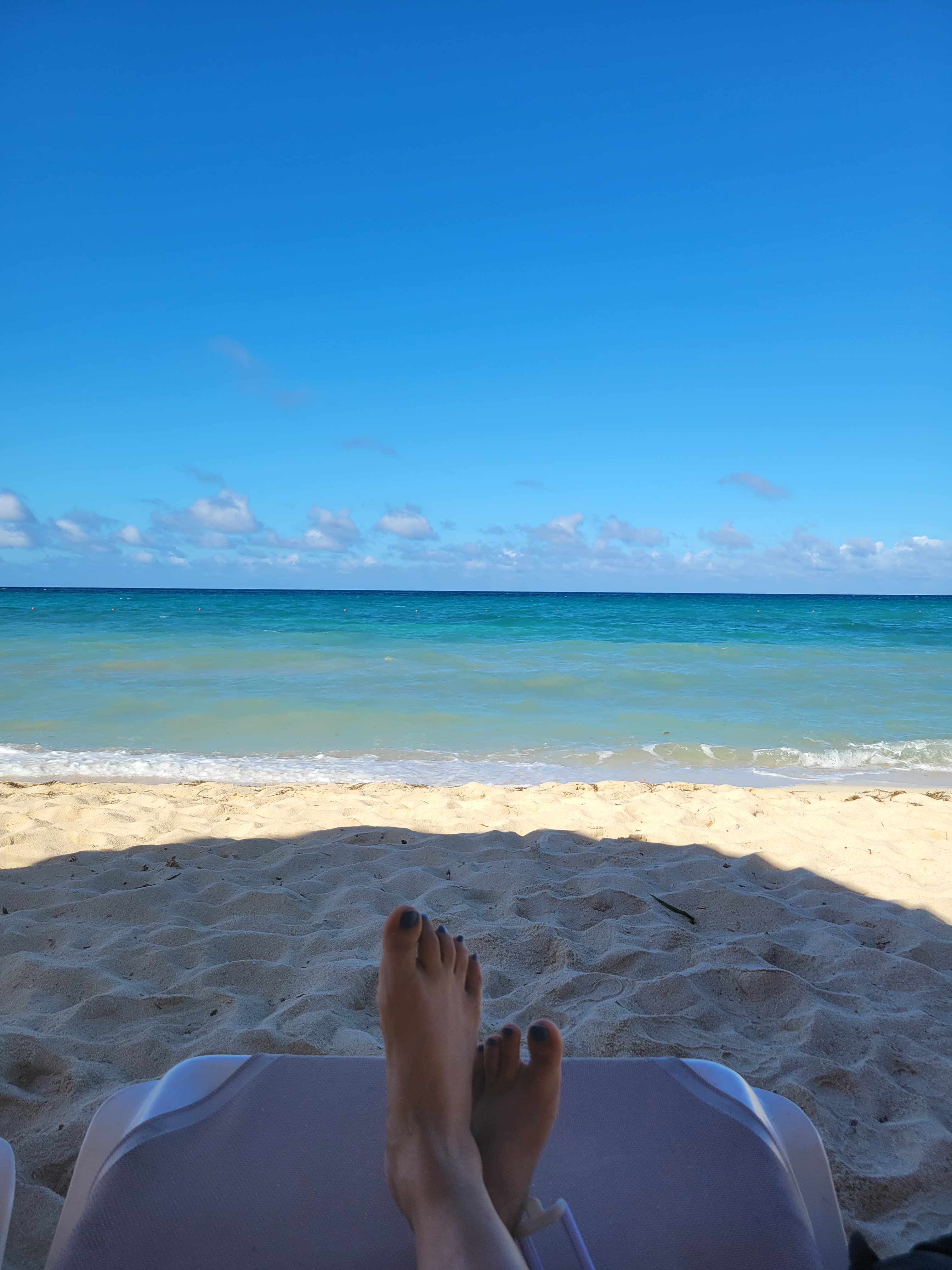 Two feet on a lounge chair looking out to a white sand beach and calm sea under sunny skies