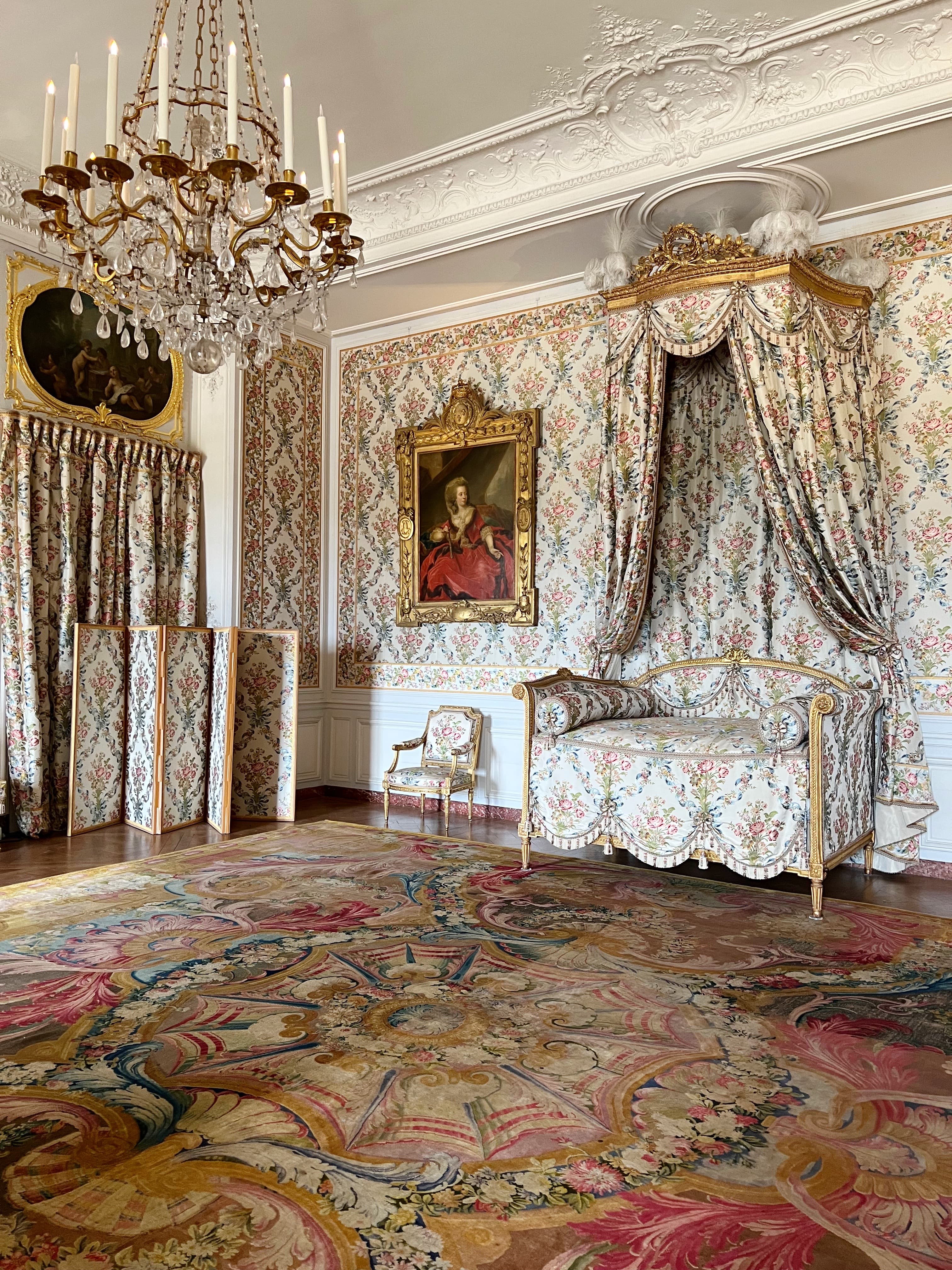 A stunning view of an old Baroque style bedroom with a gold and crystal chandelier, patterned draperies, patterned carpeting and a gold framed photo on the patterned wall. 