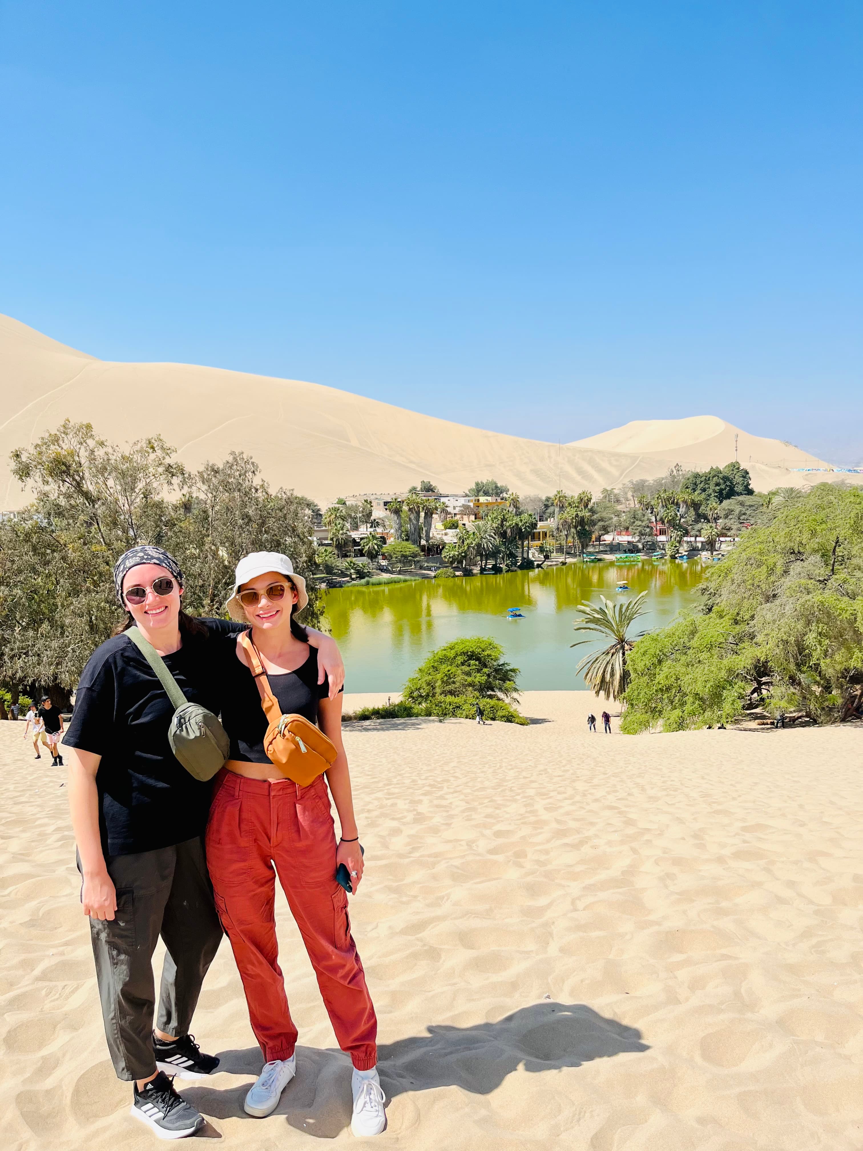 Katie and friend stand together on a sand dune, with the sun setting in the background.