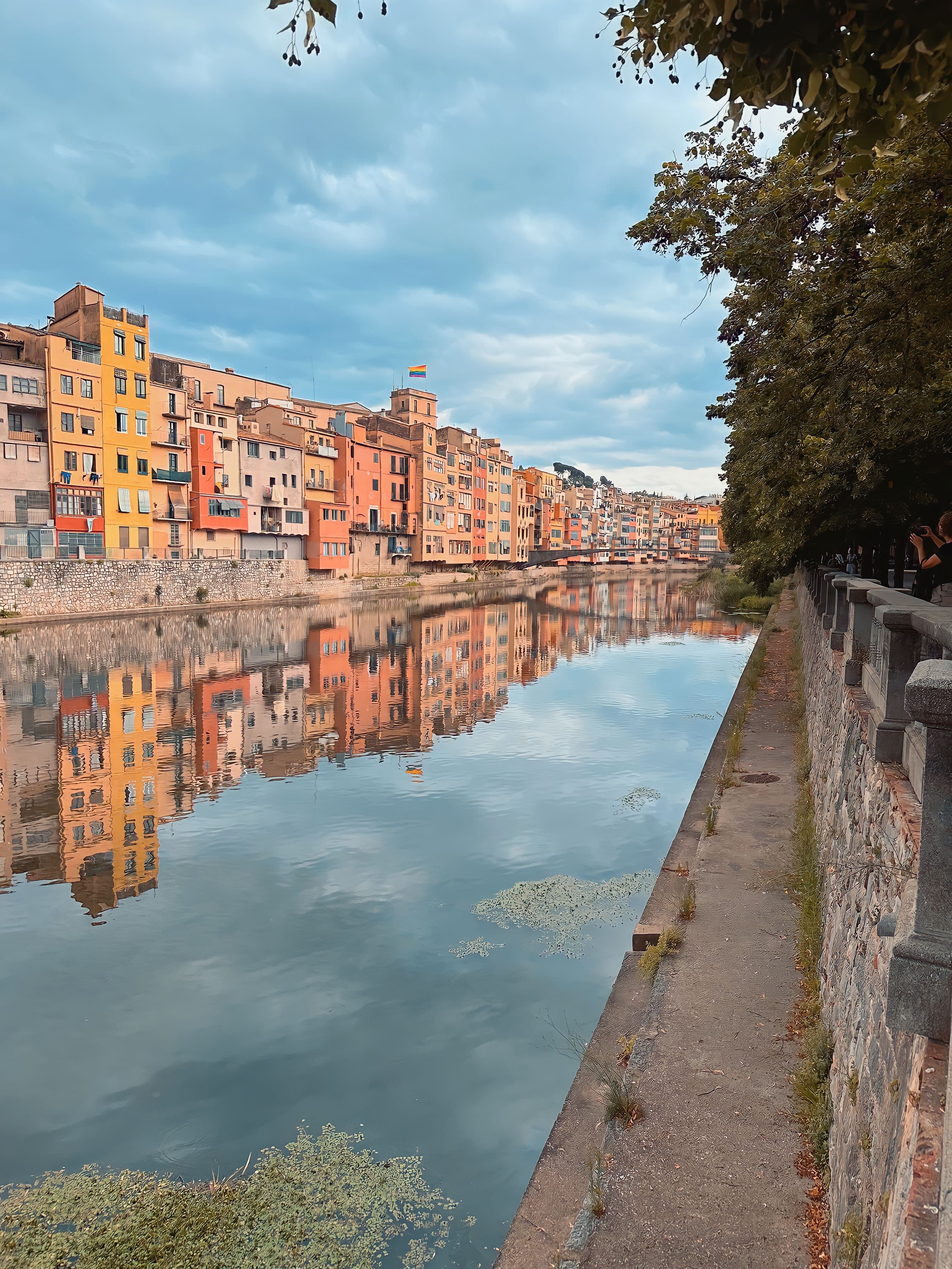 A view of tall buildings reflecting in a body of water with a brick wall and trees and partly cloudy sky.