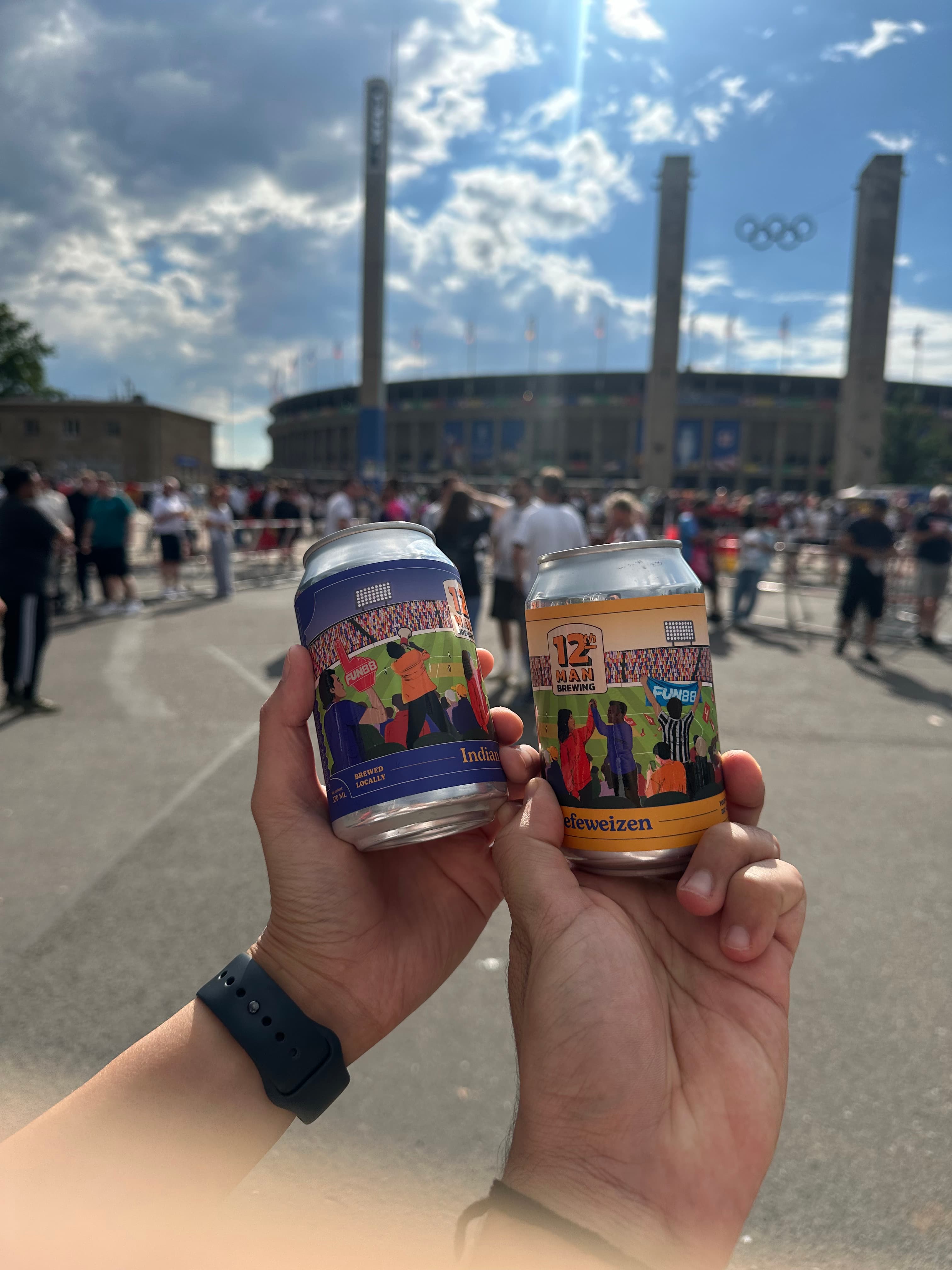 Two hands holding two colorful cans of beer with an athletic arena visible in the background