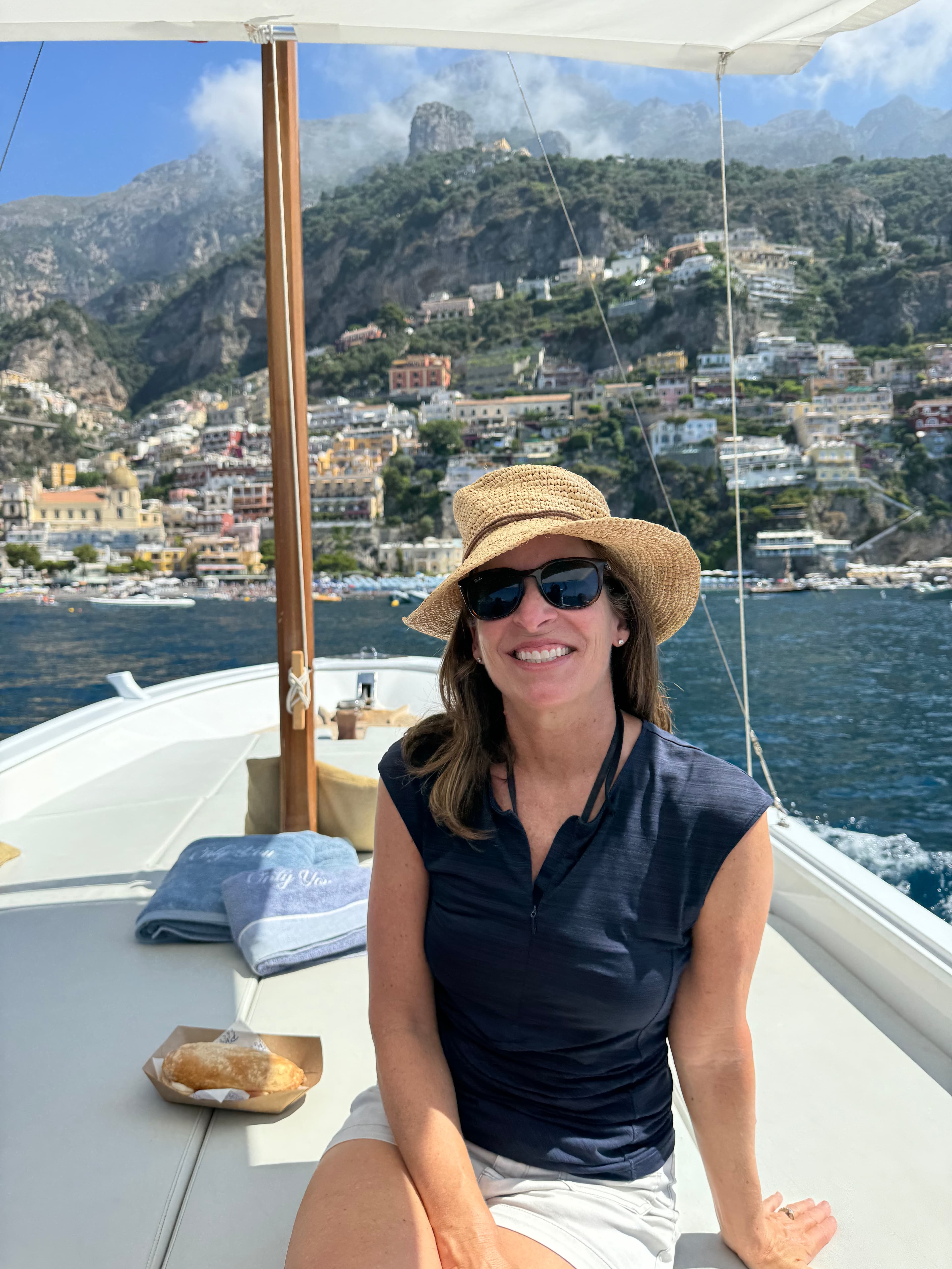 Advisor in a sun hat and sunglasses sitting on the deck of a boat with a coastal town on a hill in the background