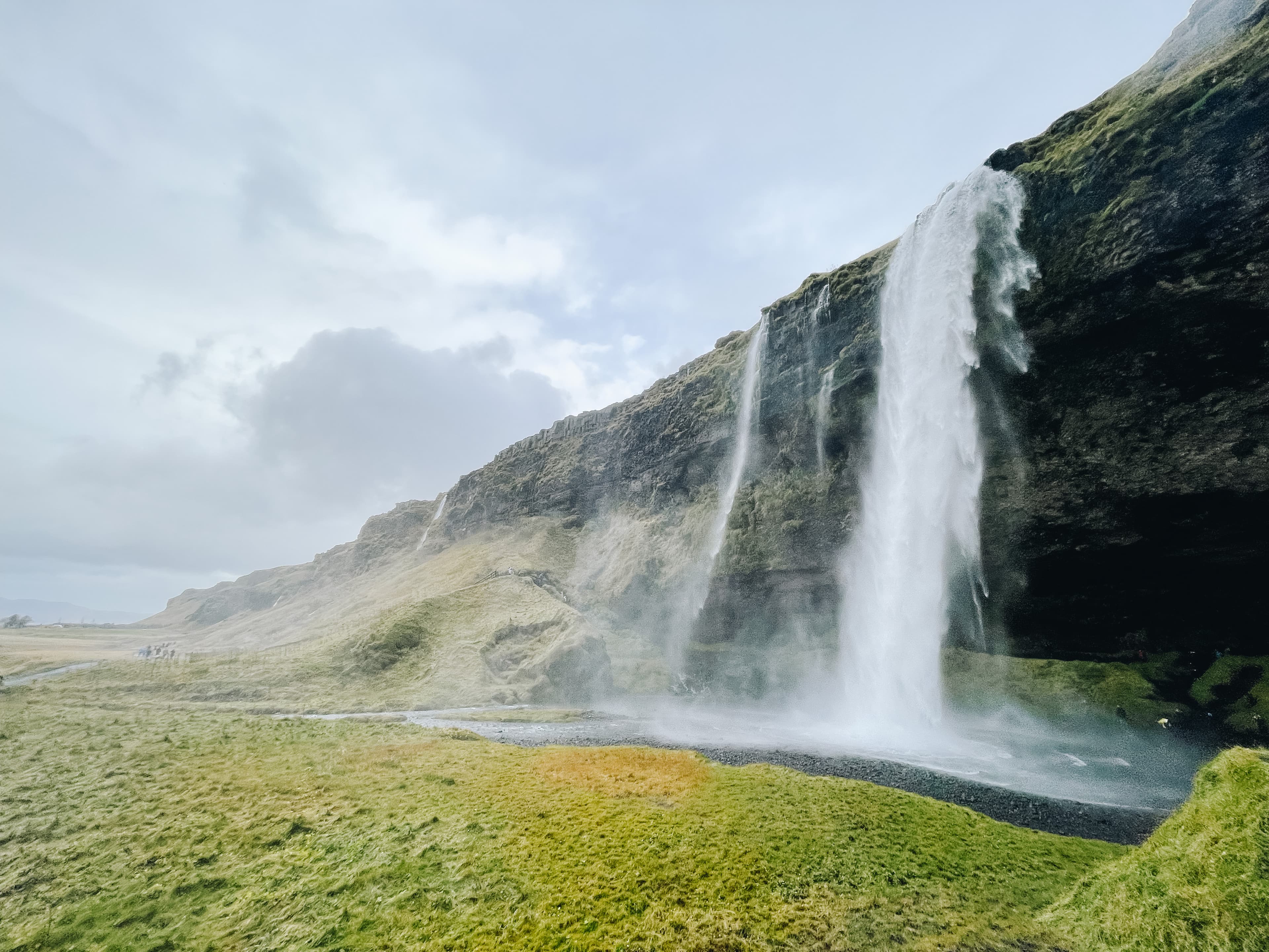 View of a remote waterfall and grassy field on a cloudy day