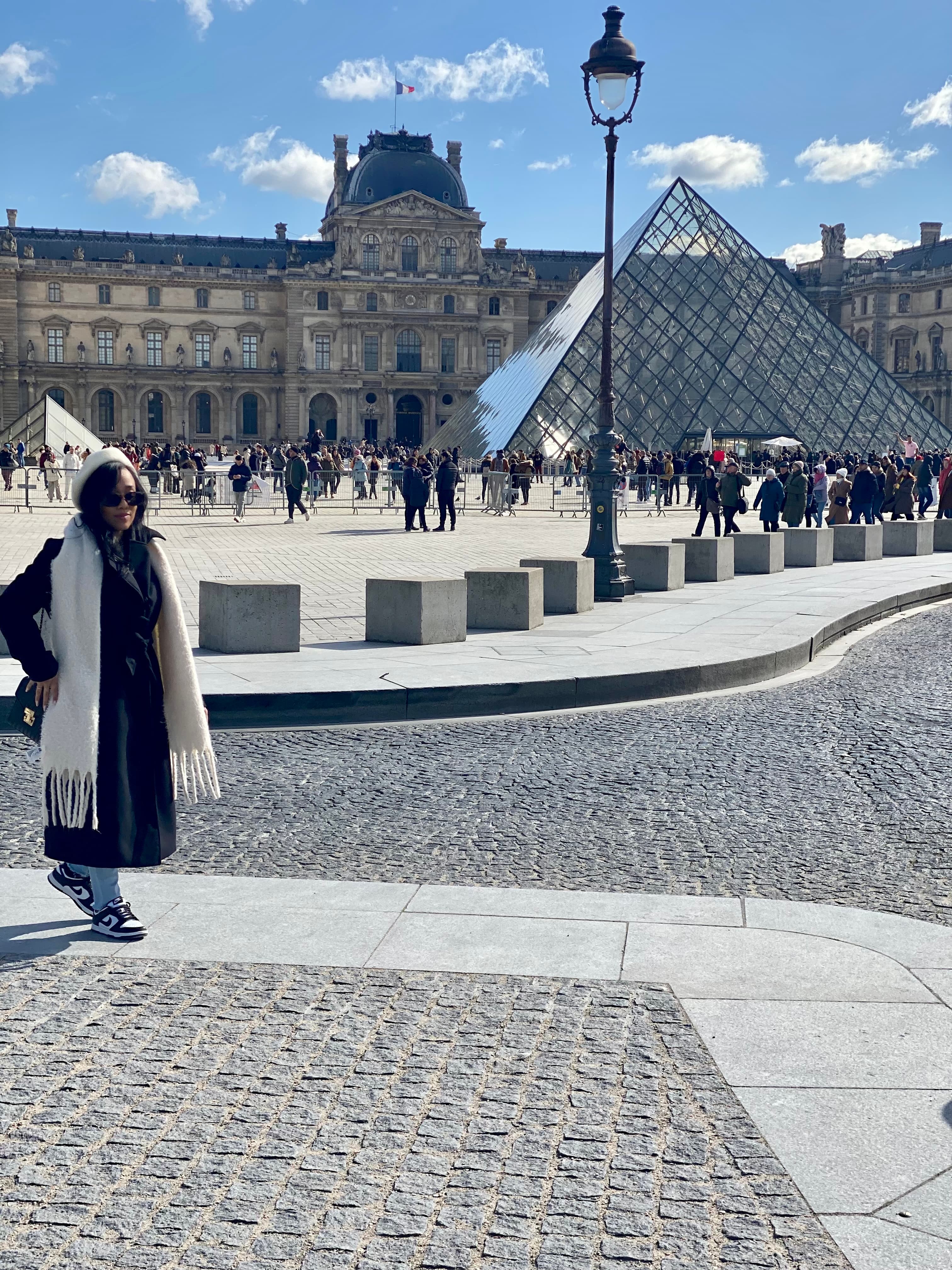 Advisor in a black jacket and white scarf posing in front of the Louvre and its glass pyramid on a sunny day