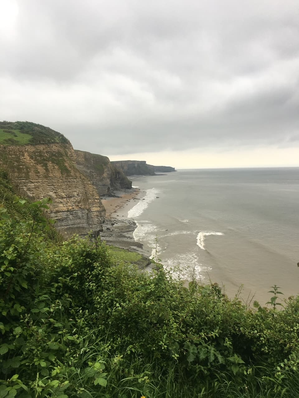 Distant view of a coastline with steep cliffs and waves breaking on shore under cloudy skies