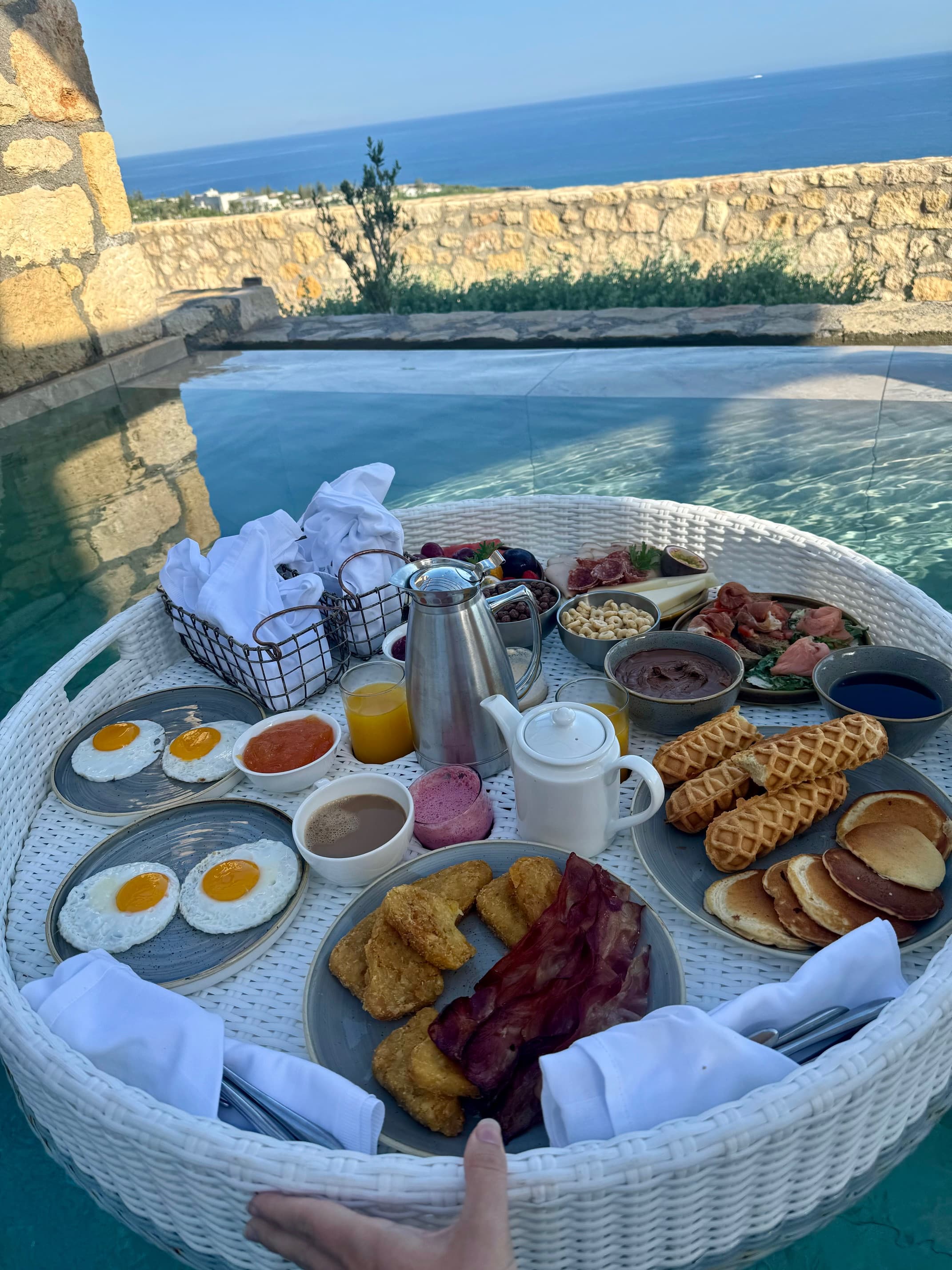A view of a breakfast spread on a white wicker tray near a swimming pool outside. It has eggs, coffee, juice and an assortment of pastries. 