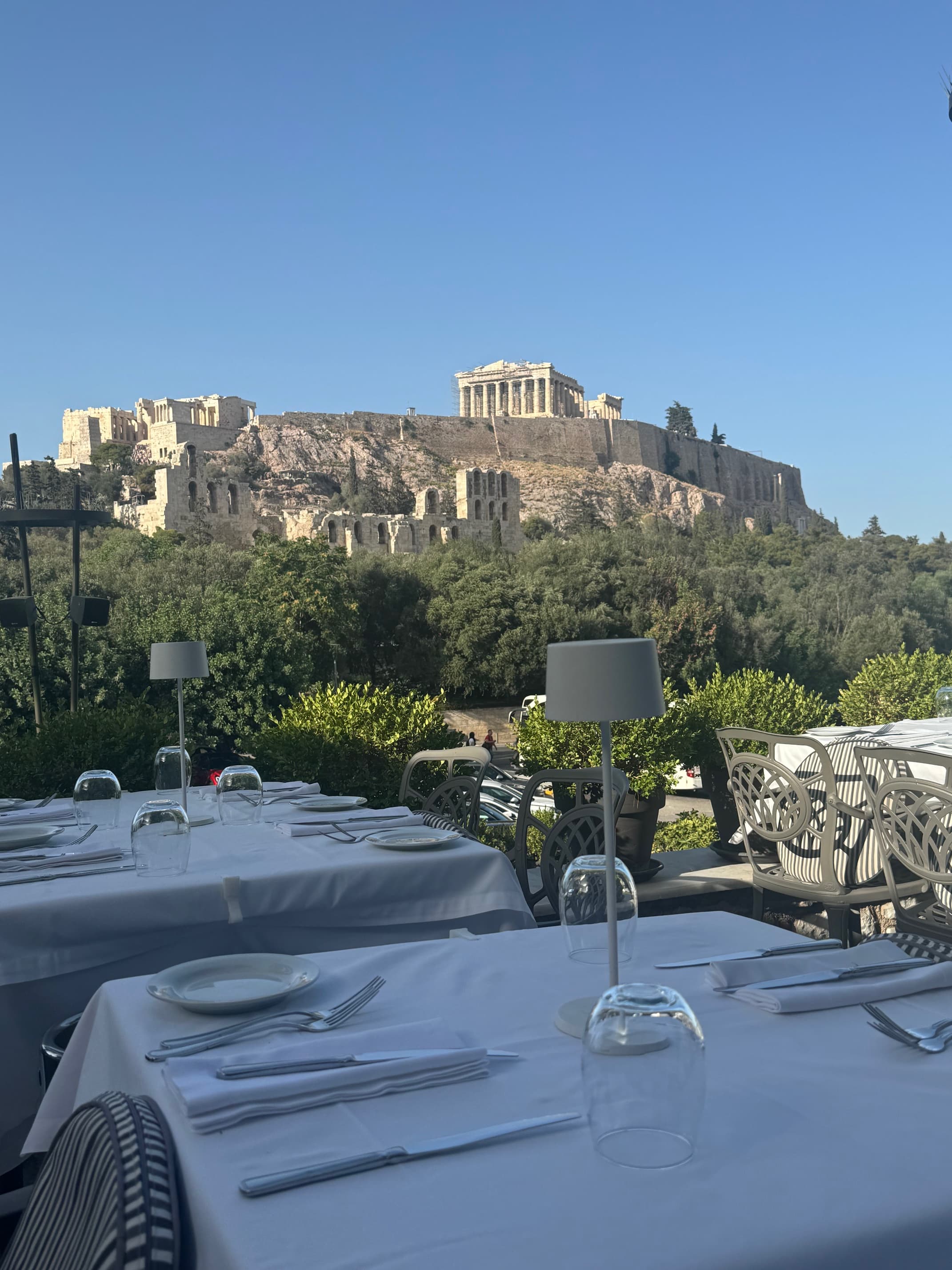 An image of a dining table outside with place settings and a view of Greece in the background on a beautiful day. 