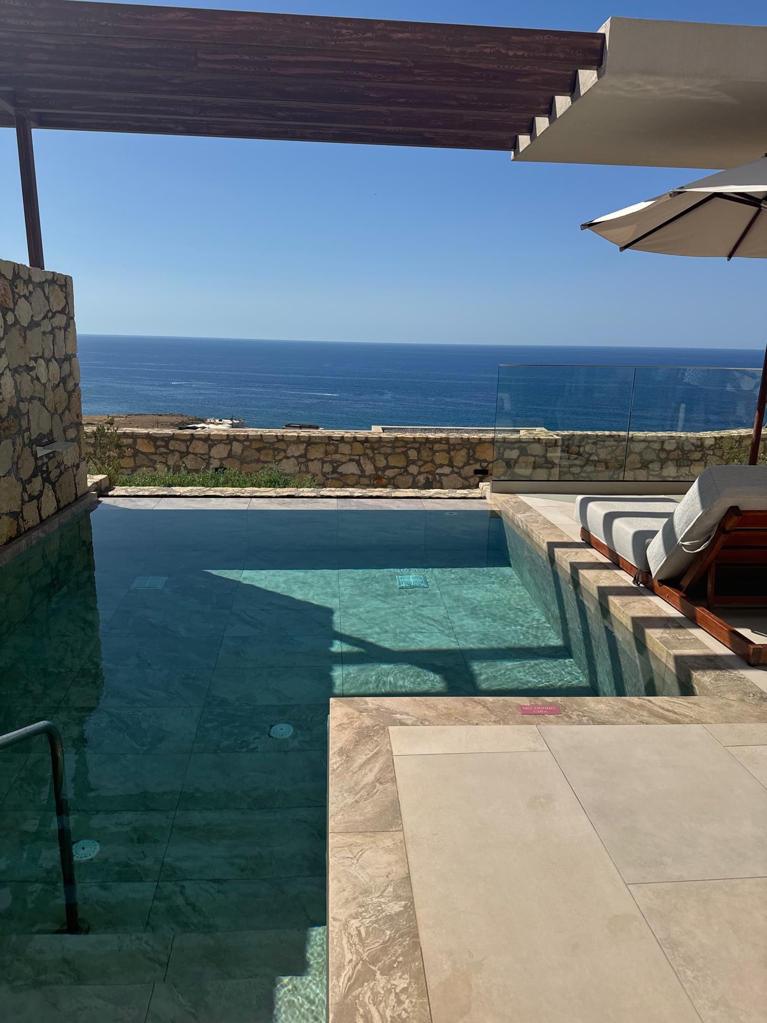 A picture of a swimming pool outside with a stone ledge, umbrella, lounge chair and view of the sea in the distance. 