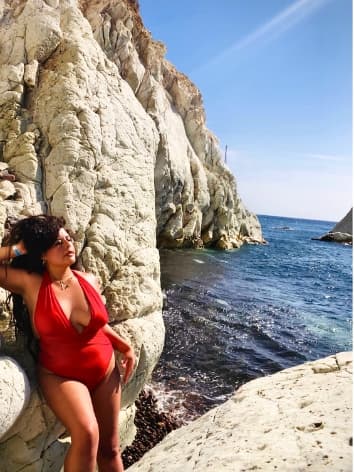 Advisor in a red one-piece bathing suit leaning against a cliffside by the sea on a sunny day
