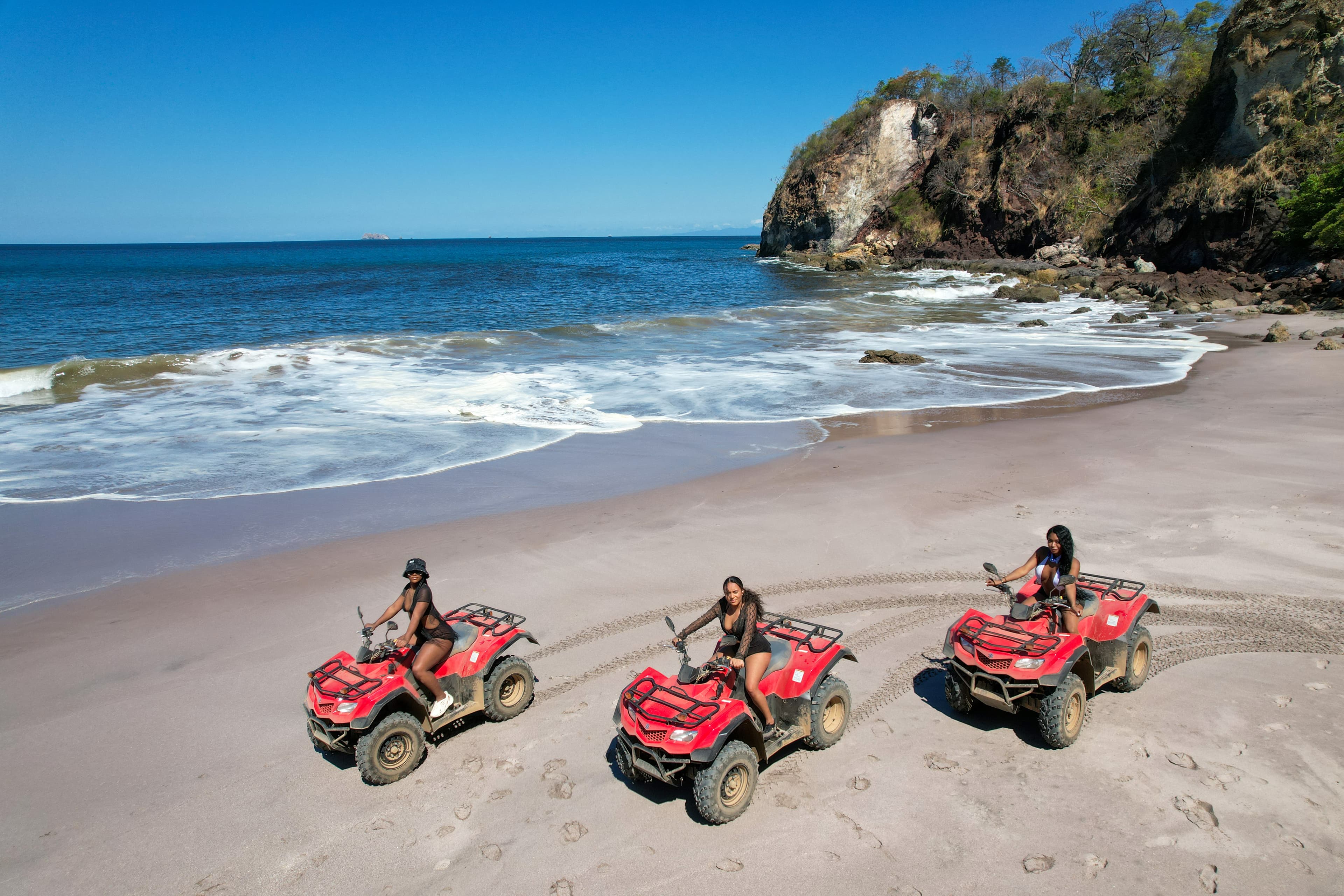 View of three people riding red four-wheel vehicles on a wide stretch of beach lined with cliffs on a clear day