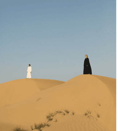Two people, one in all black and one in all white, standing atop desert sand dunes under clear skies