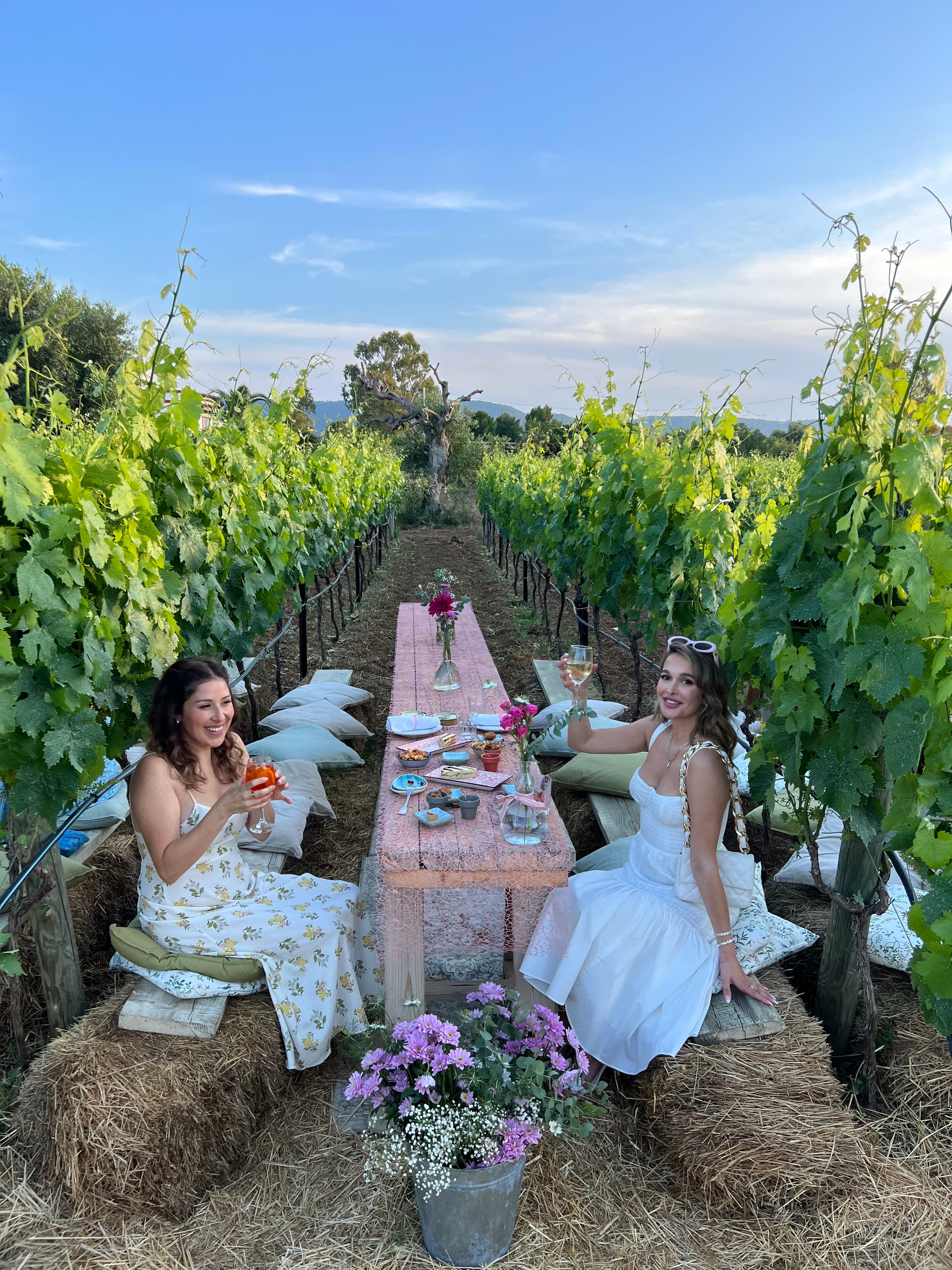 Advisor and another woman in white dresses sitting at a long table set in between stalks of corn on a sunny day